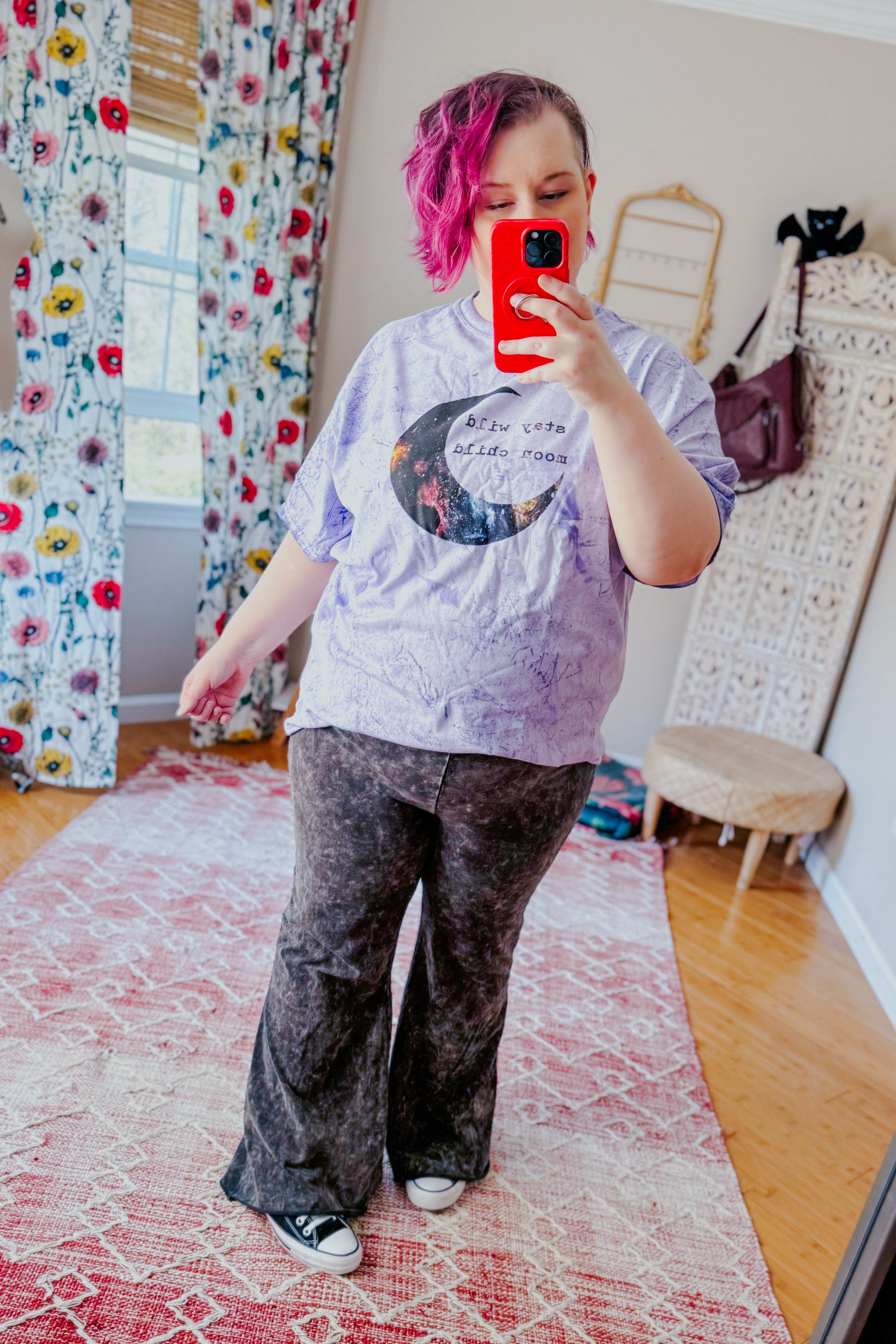 Woman with pink hair wearing purple moon graphic tee and dark wide-leg pants taking a mirror selfie indoors