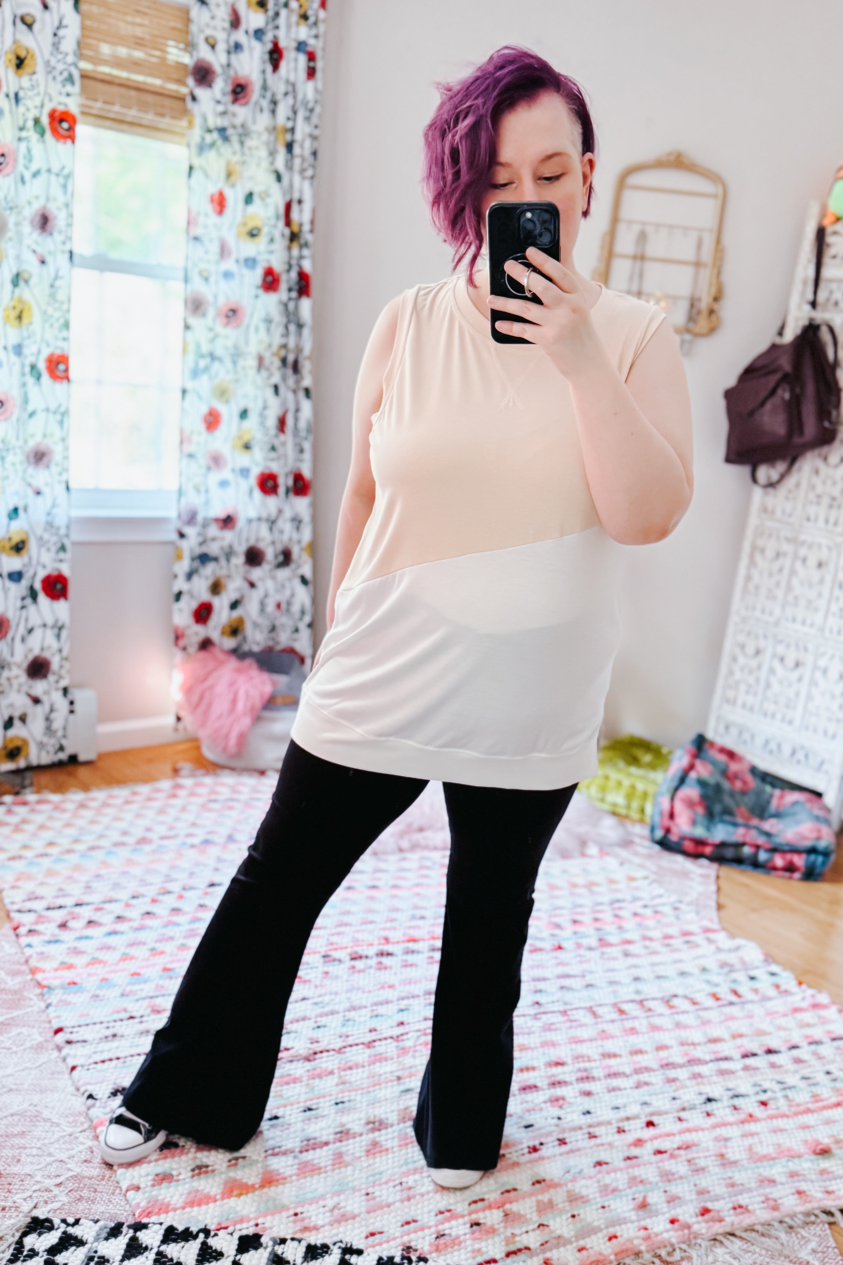 Woman with purple hair wearing a cream color block sleeveless top paired with black pants in a bright eclectic room