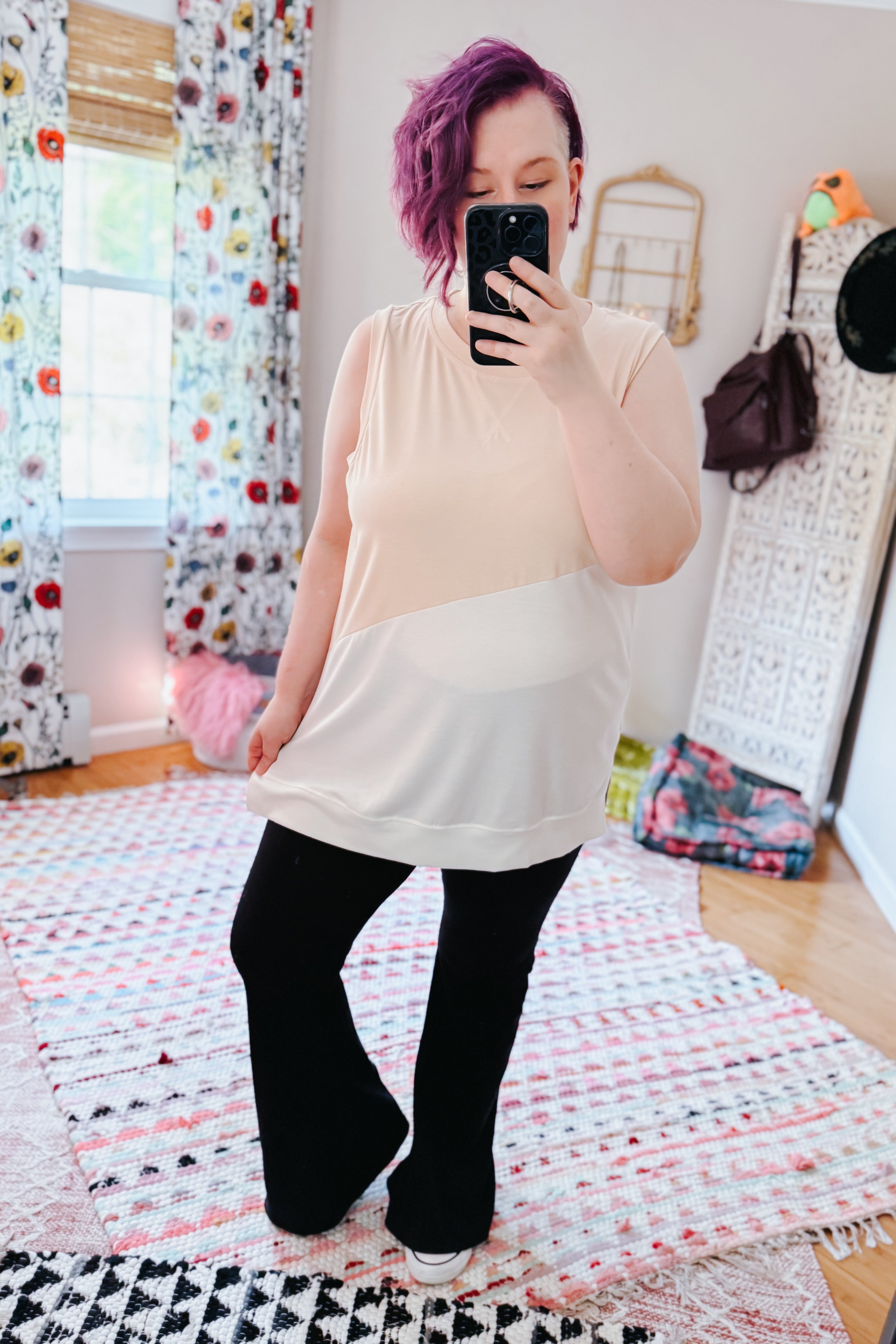 Woman with purple hair wearing a cream color block sleeveless top and black pants in a cozy eclectic room