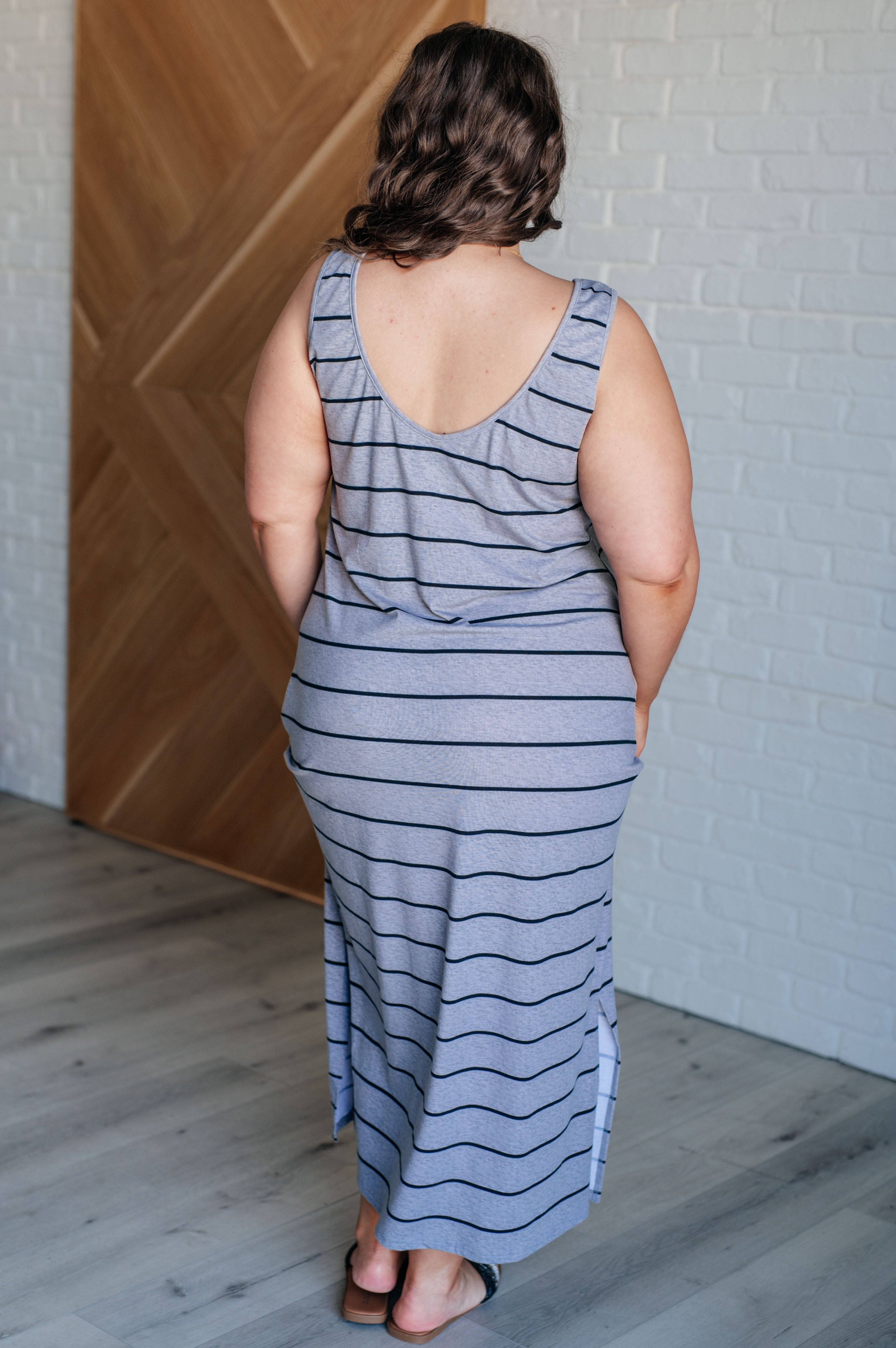 Curvy model wearing a sleeveless gray striped maxi dress with a scoop back standing indoors by wooden door and white brick wall