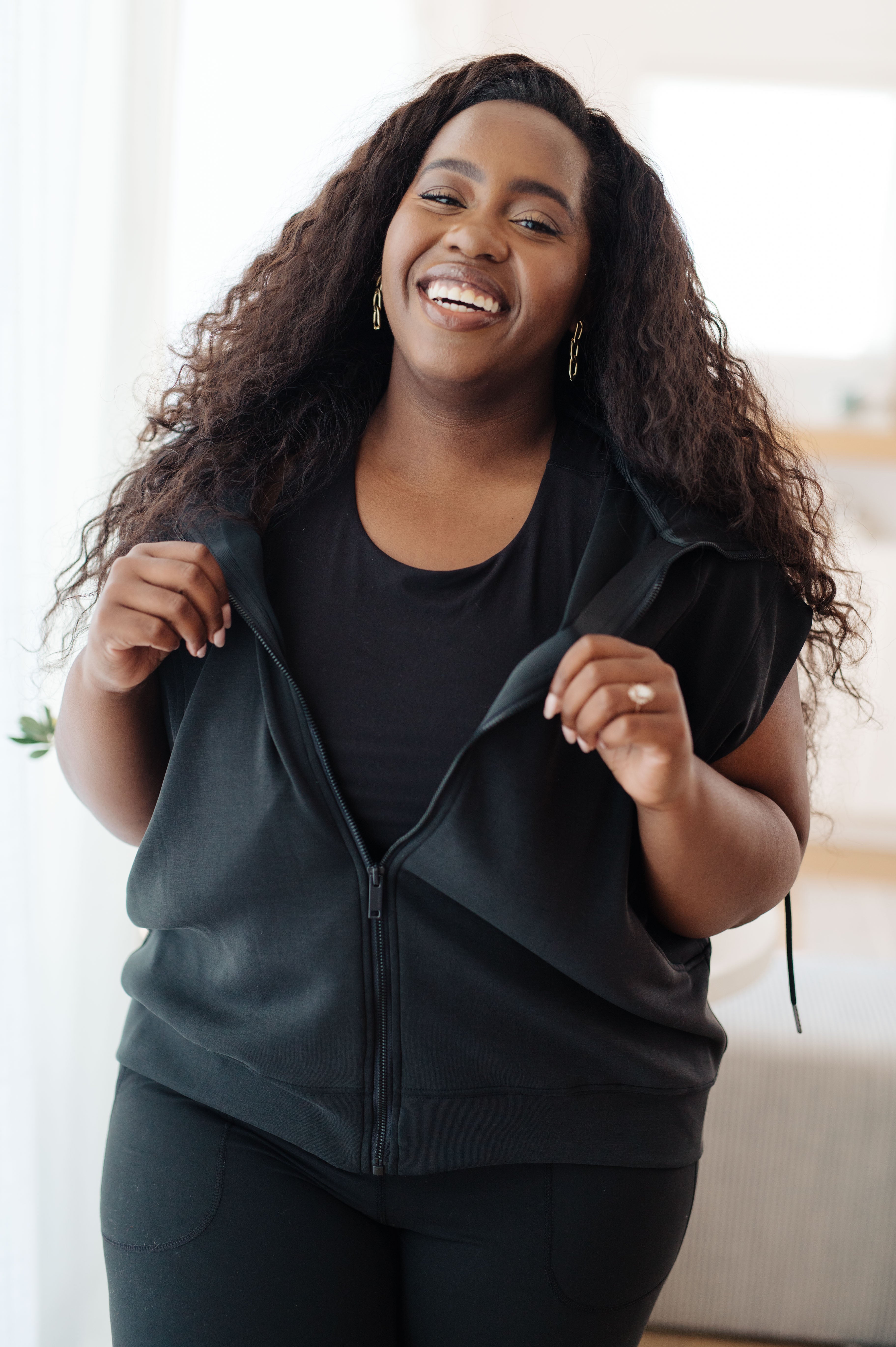 Smiling woman wearing a black sleeveless hoodie unzipped over a black top in a bright indoor setting