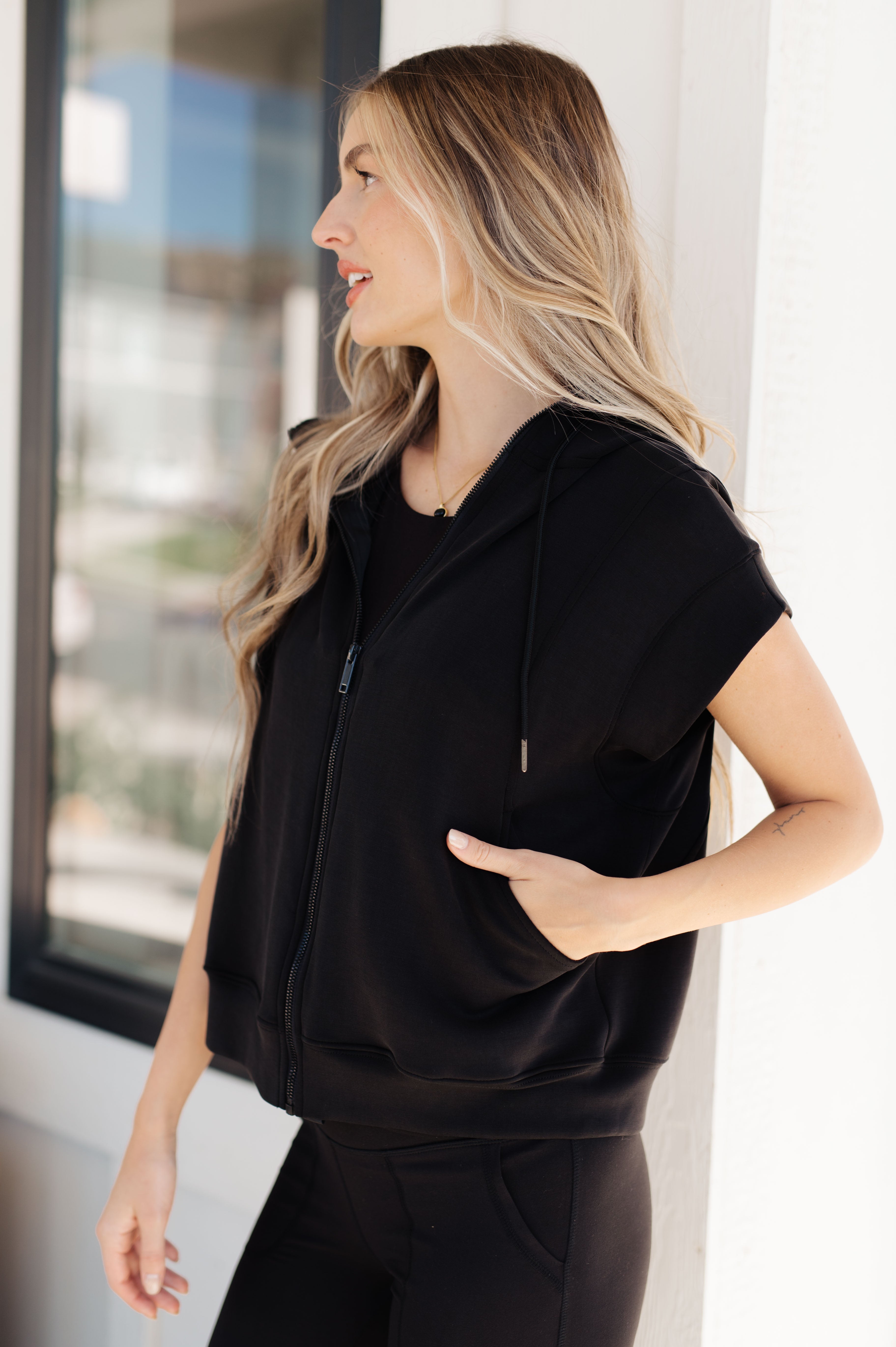 Woman wearing black sleeveless hoodie with zipper and hood standing outdoors by window in casual style