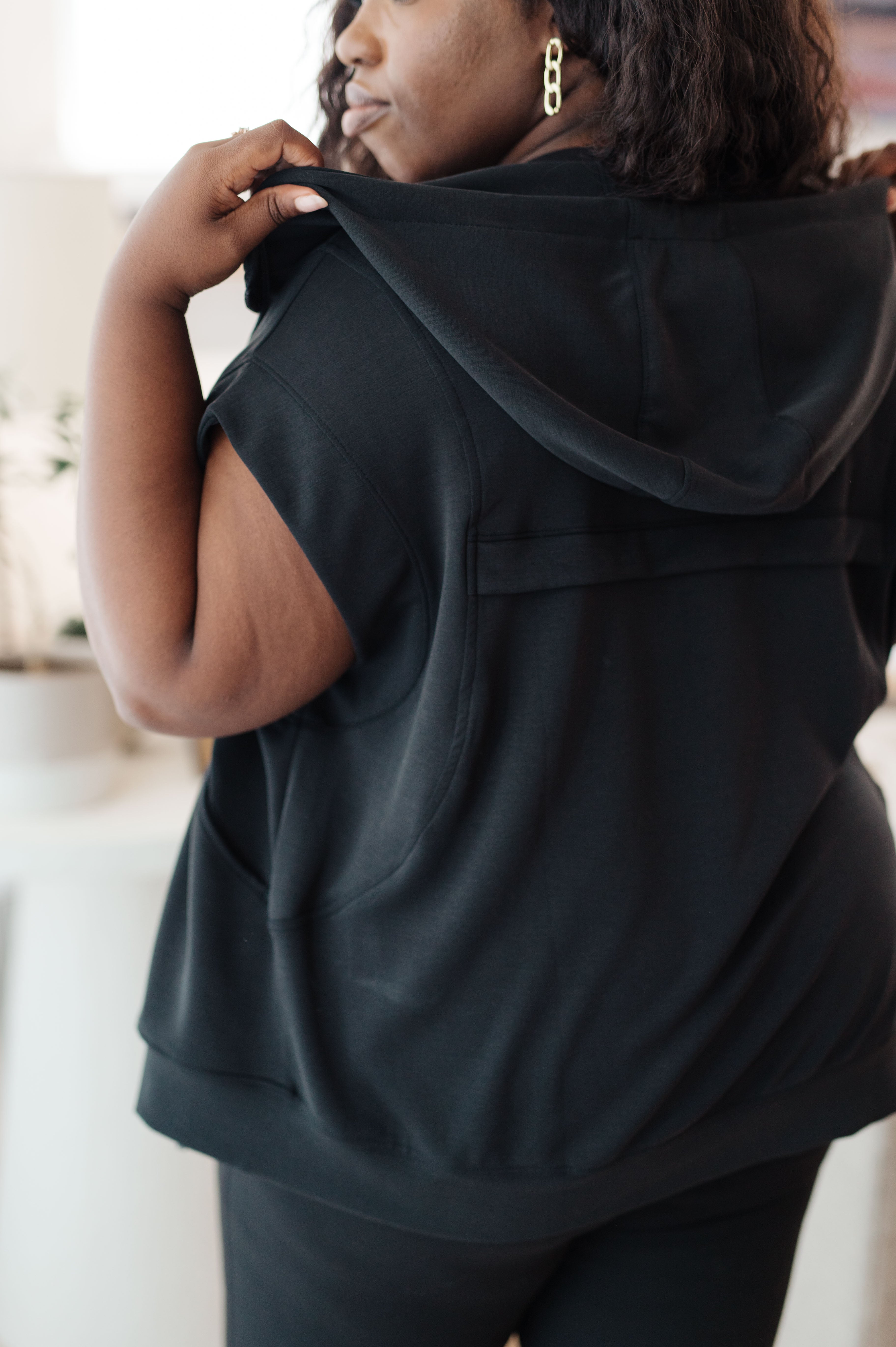 Curvy woman wearing black sleeveless hoodie with a hood pulled up, accessorized with gold chain earrings indoors