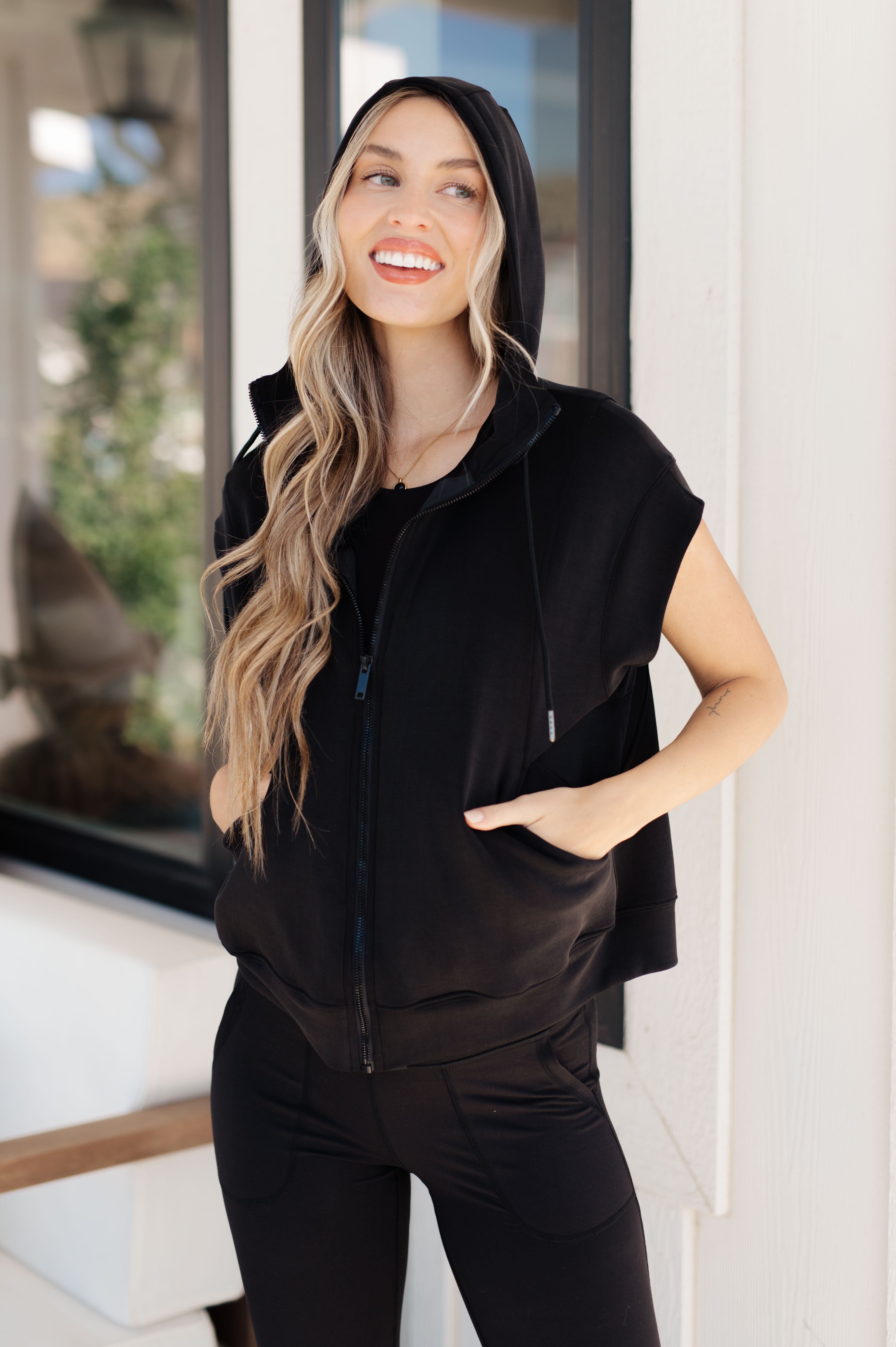 Smiling woman with long wavy hair wearing a black sleeveless zip-up hoodie with hood outdoors by white wall