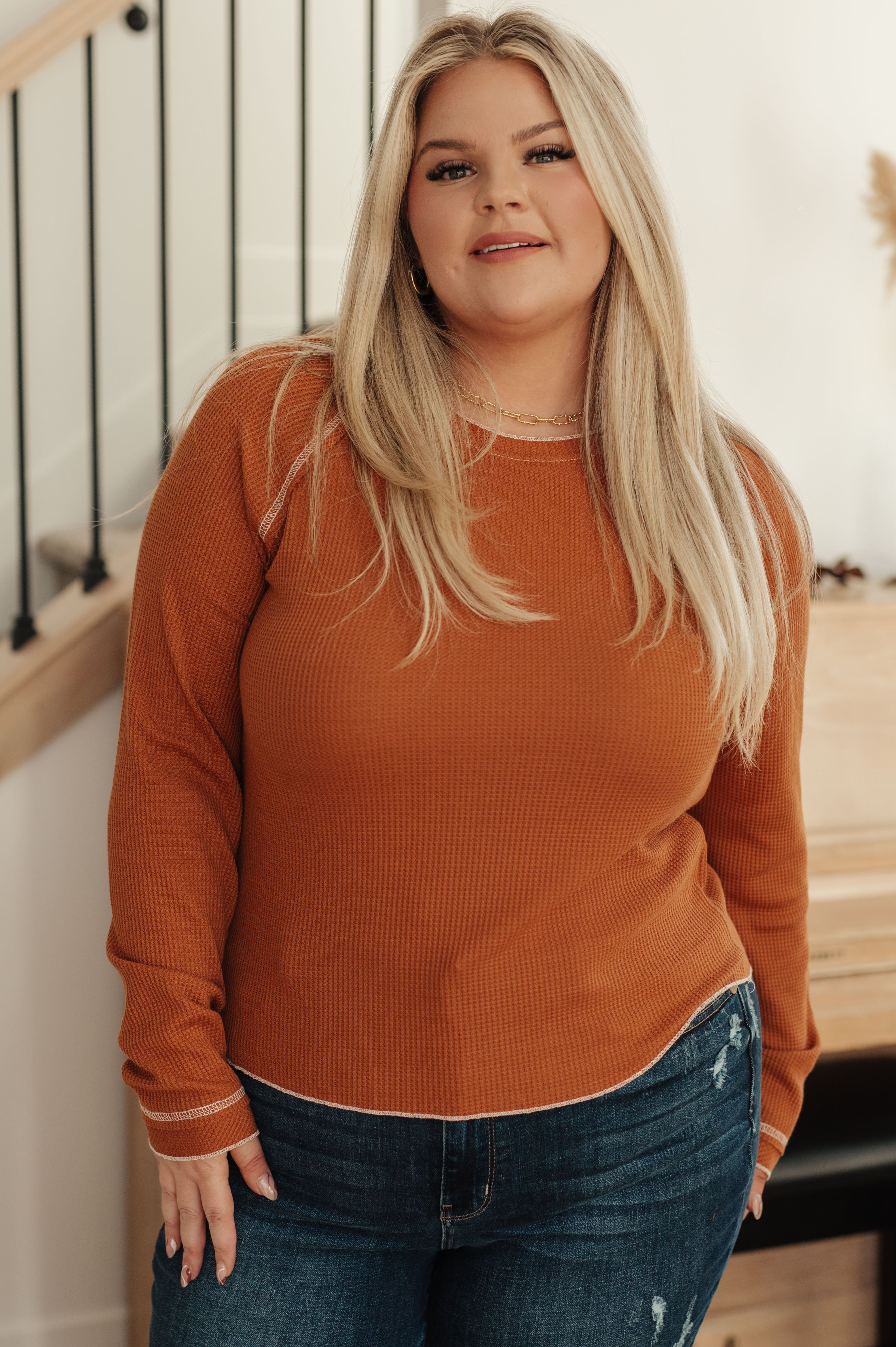 Curvy woman wearing rust-colored waffle knit long sleeve top with white stitch details, standing indoors by staircase railing