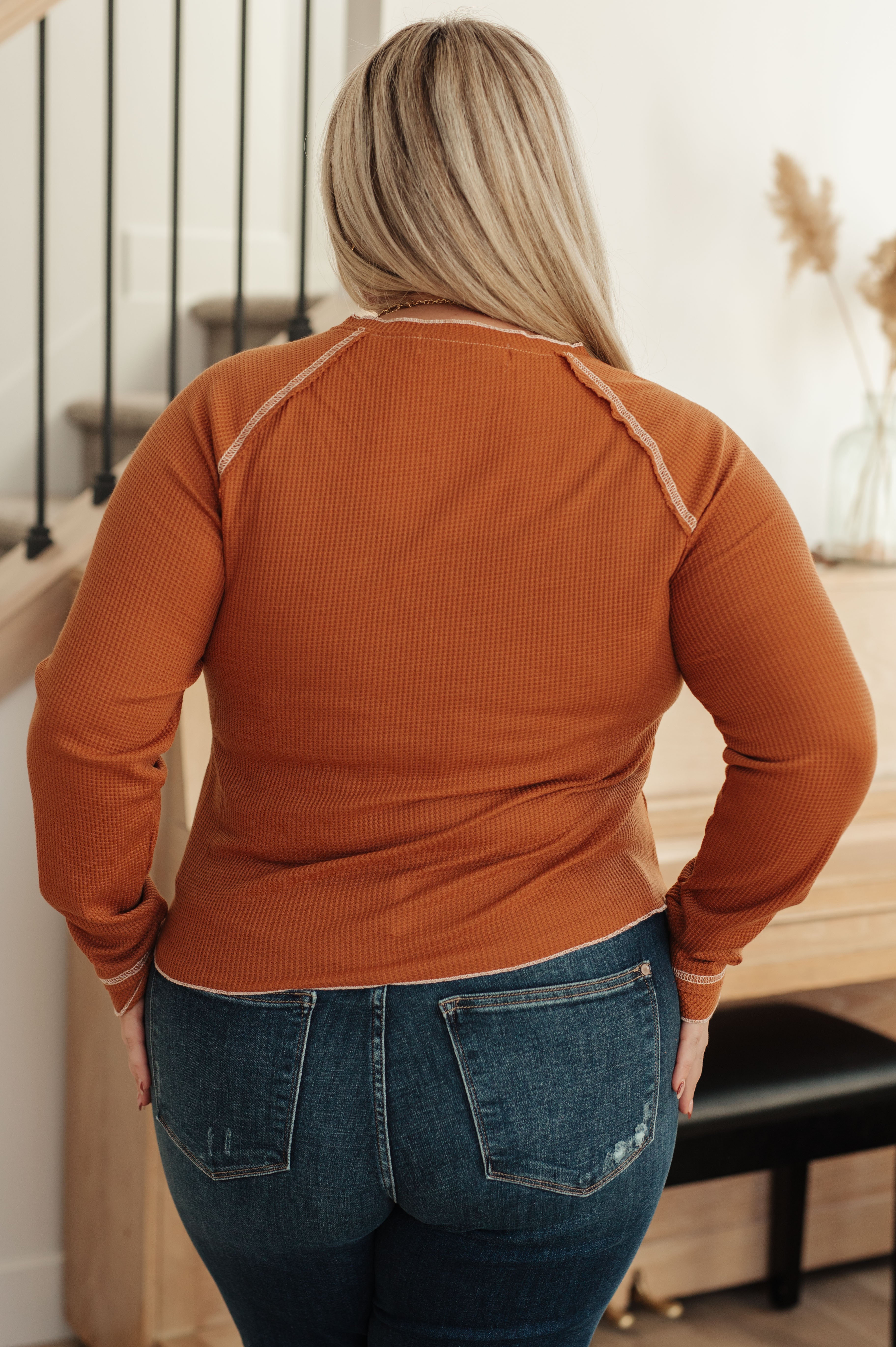 Plus-size woman wearing rust waffle knit top and blue jeans standing indoors near wooden piano and staircase railing