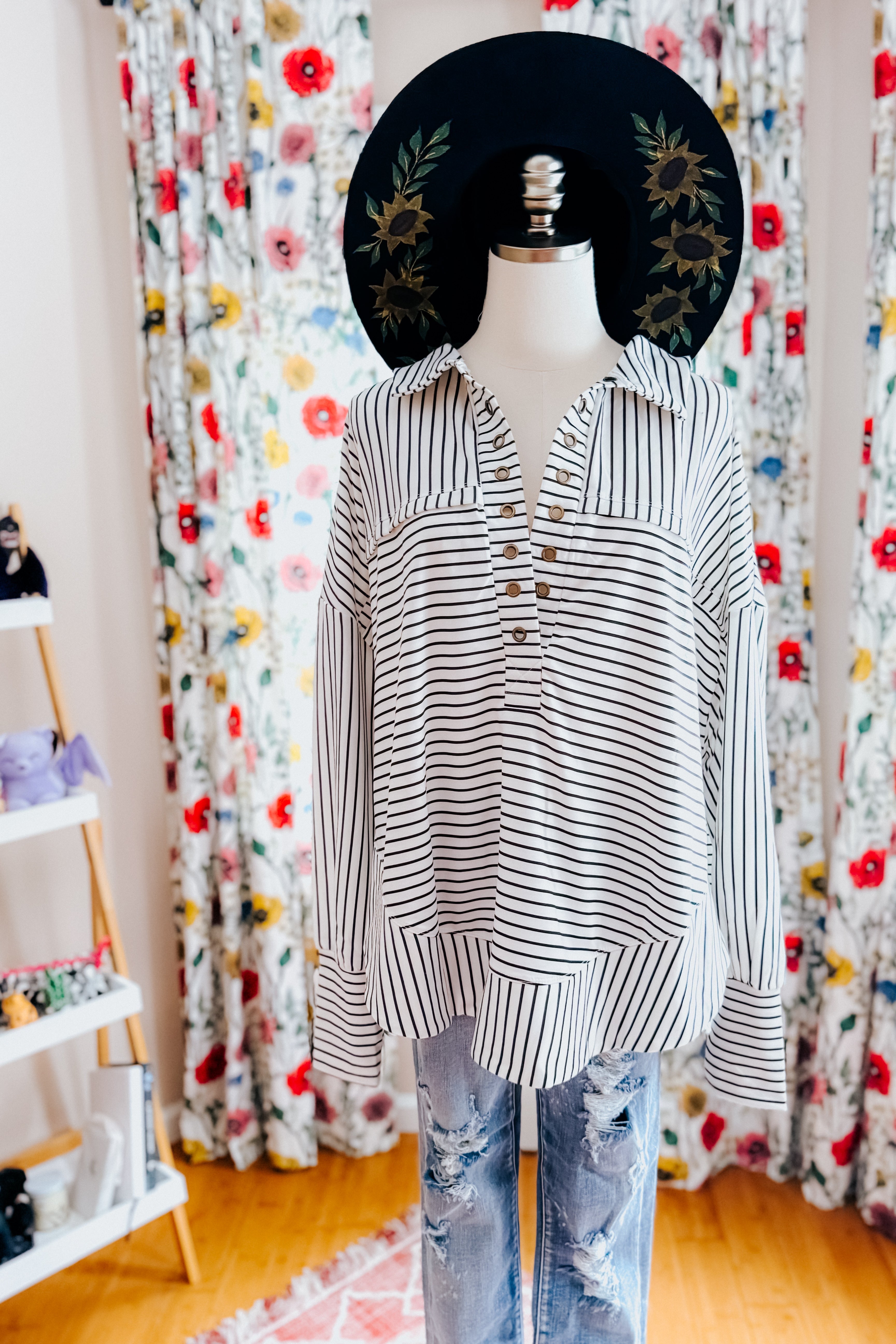 A dress form wearing by a black and white striped long-sleeve collared pullover, a black hand painted wide brim hat and blue jeans in an indoor setting.