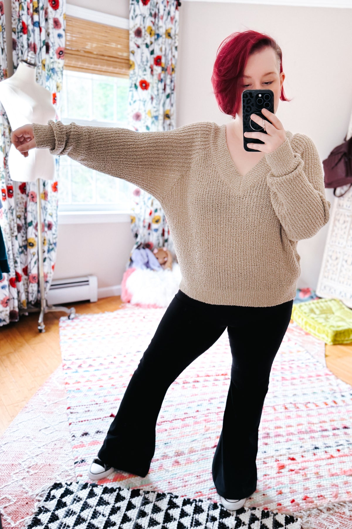 Red haired woman taking a bedroom mirror selfie in a beige V-neck sweater with relaxed sleeves black flared pants and white sneakers