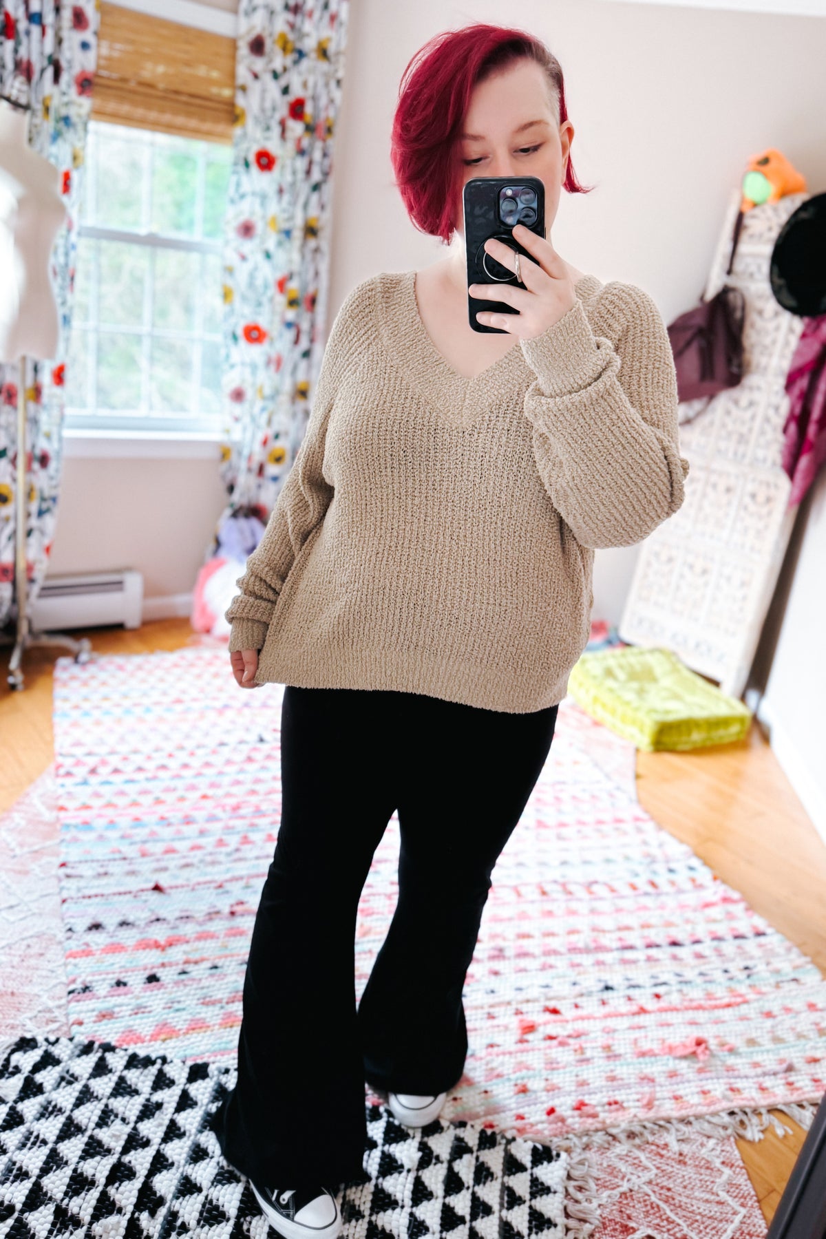 Red haired woman taking a bedroom mirror selfie in a beige V neck sweater with black pants and sneakers on colorful layered rugs