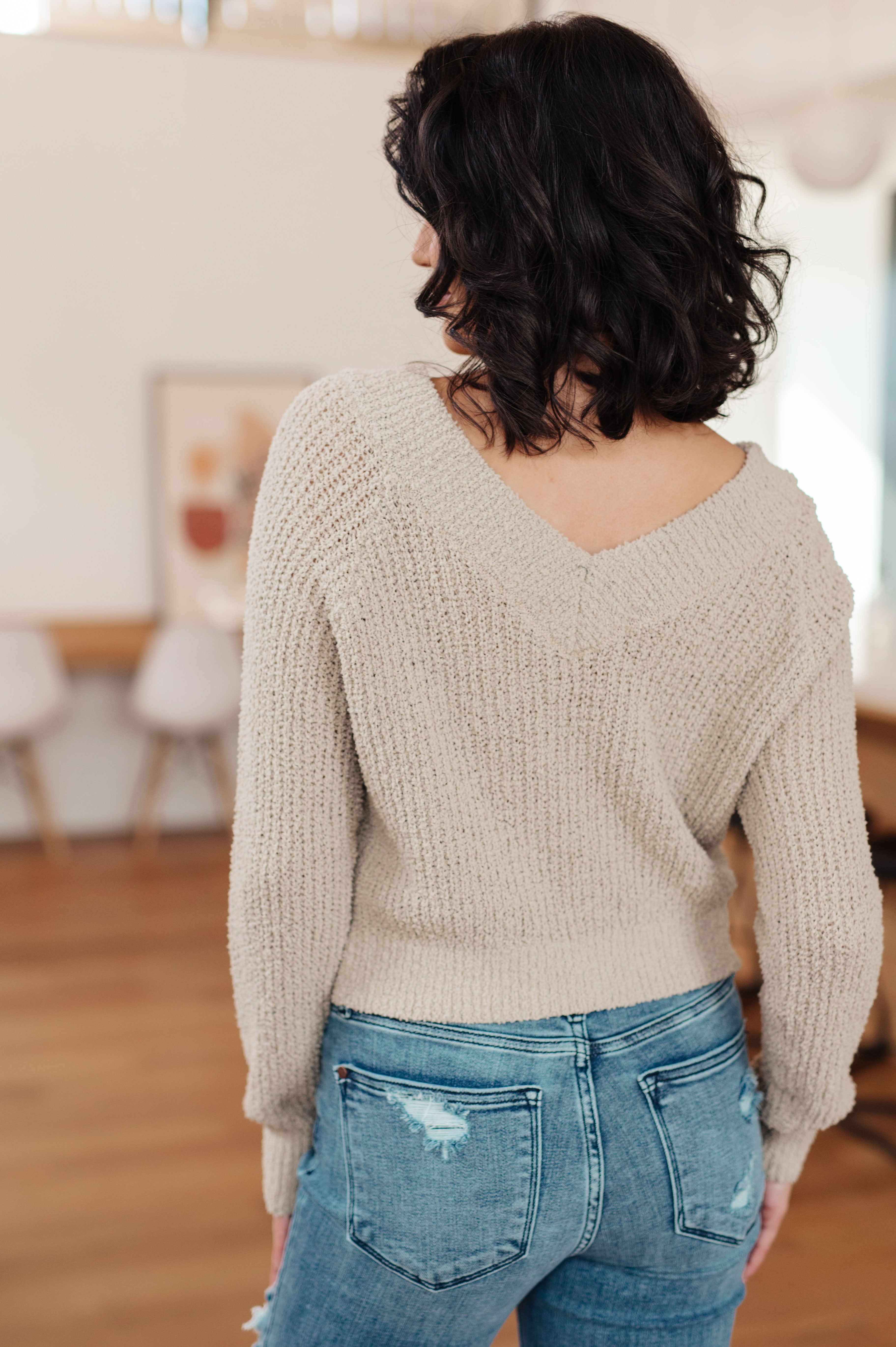 Woman wearing beige textured V-neck sweater with long sleeves and ripped blue jeans seen from back indoors