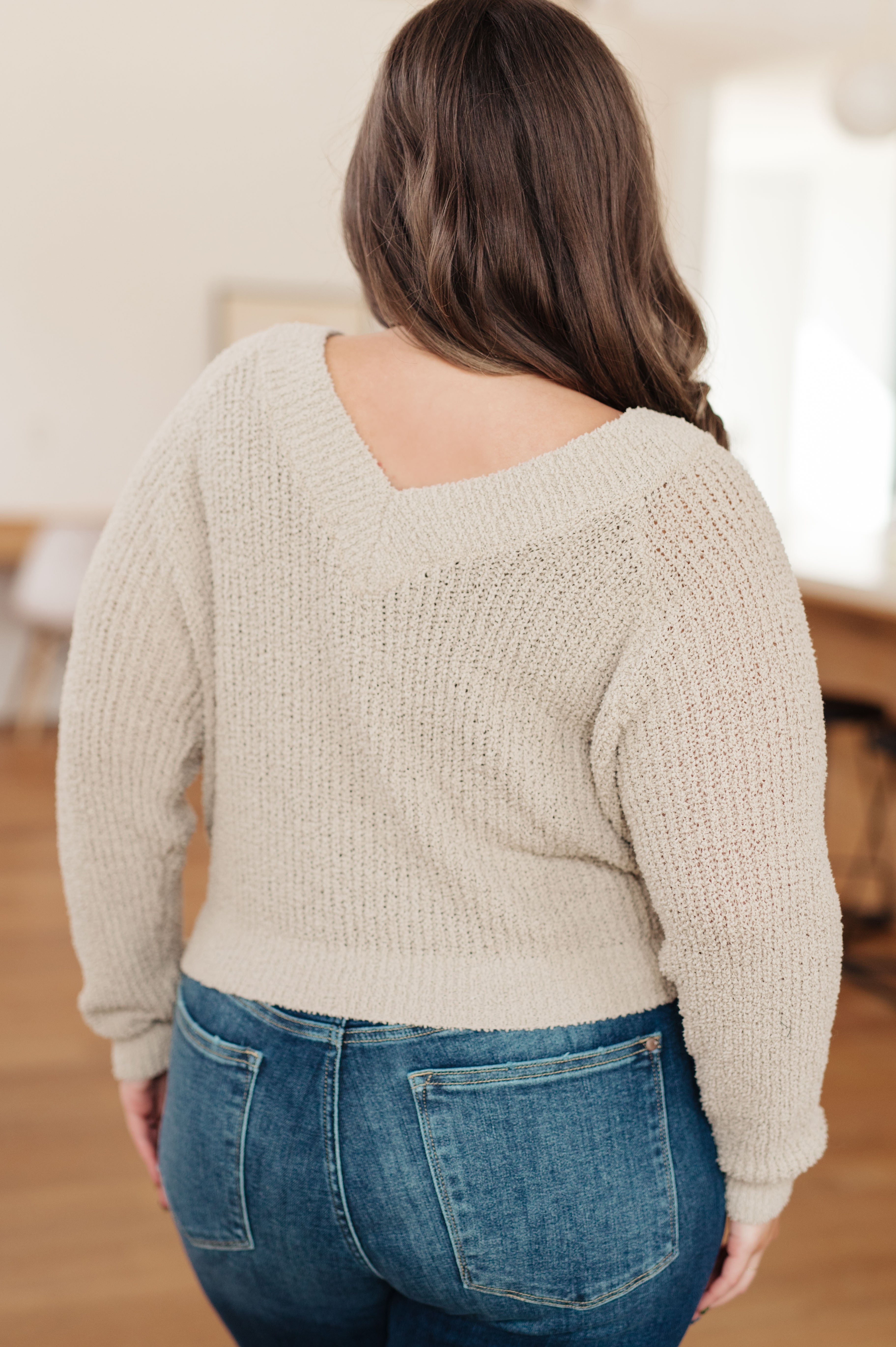 Curvy woman wearing beige textured knit sweater with deep V-back paired with blue jeans in cozy indoor setting