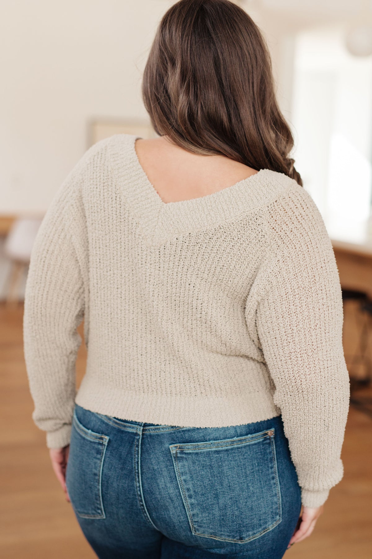 Back view of a woman wearing a beige knit sweater with a V neckline and blue jeans in a warm home interior