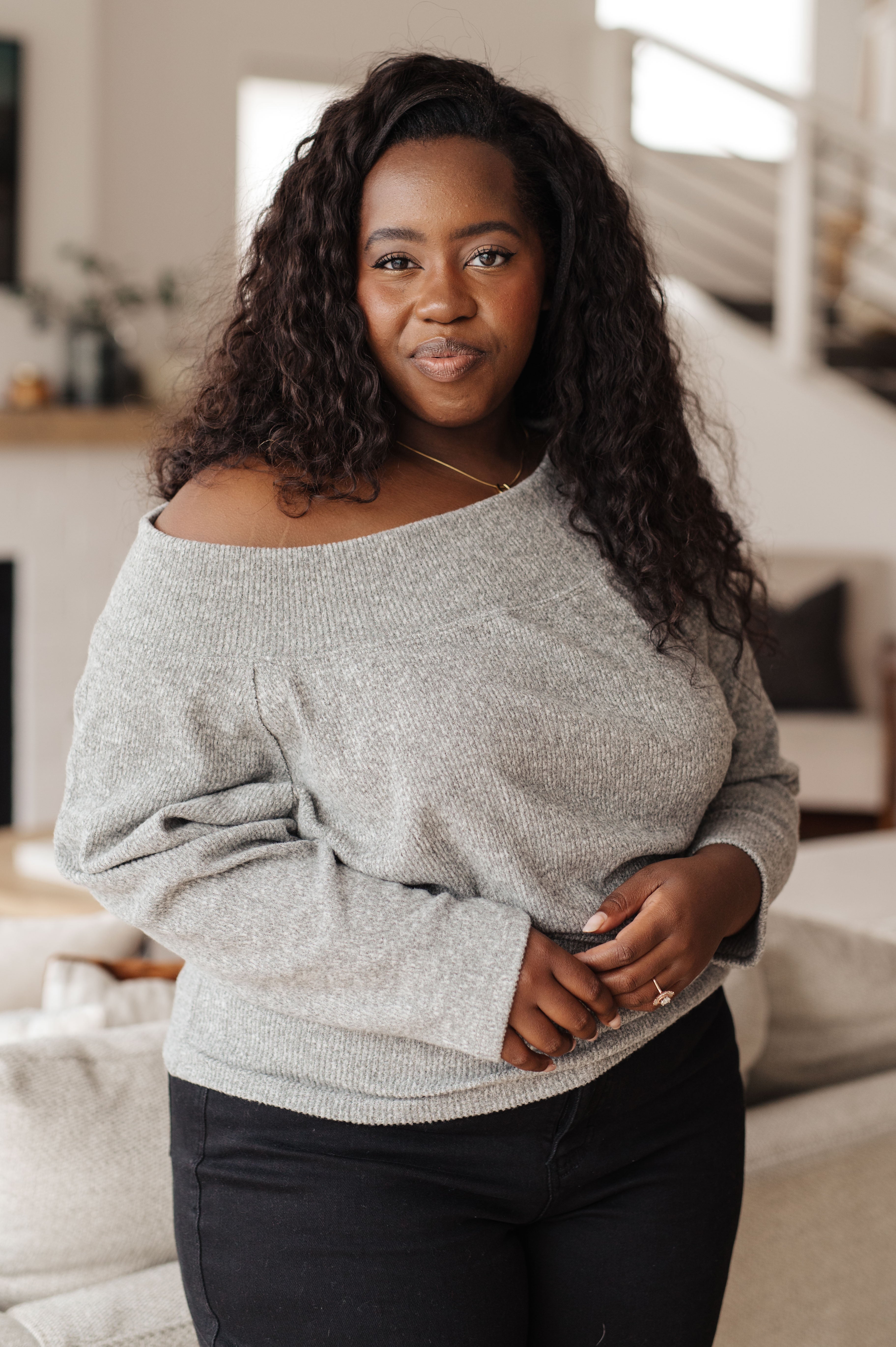 Confident woman wearing grey knit long sleeve top with off-shoulder detail in relaxed indoor setting