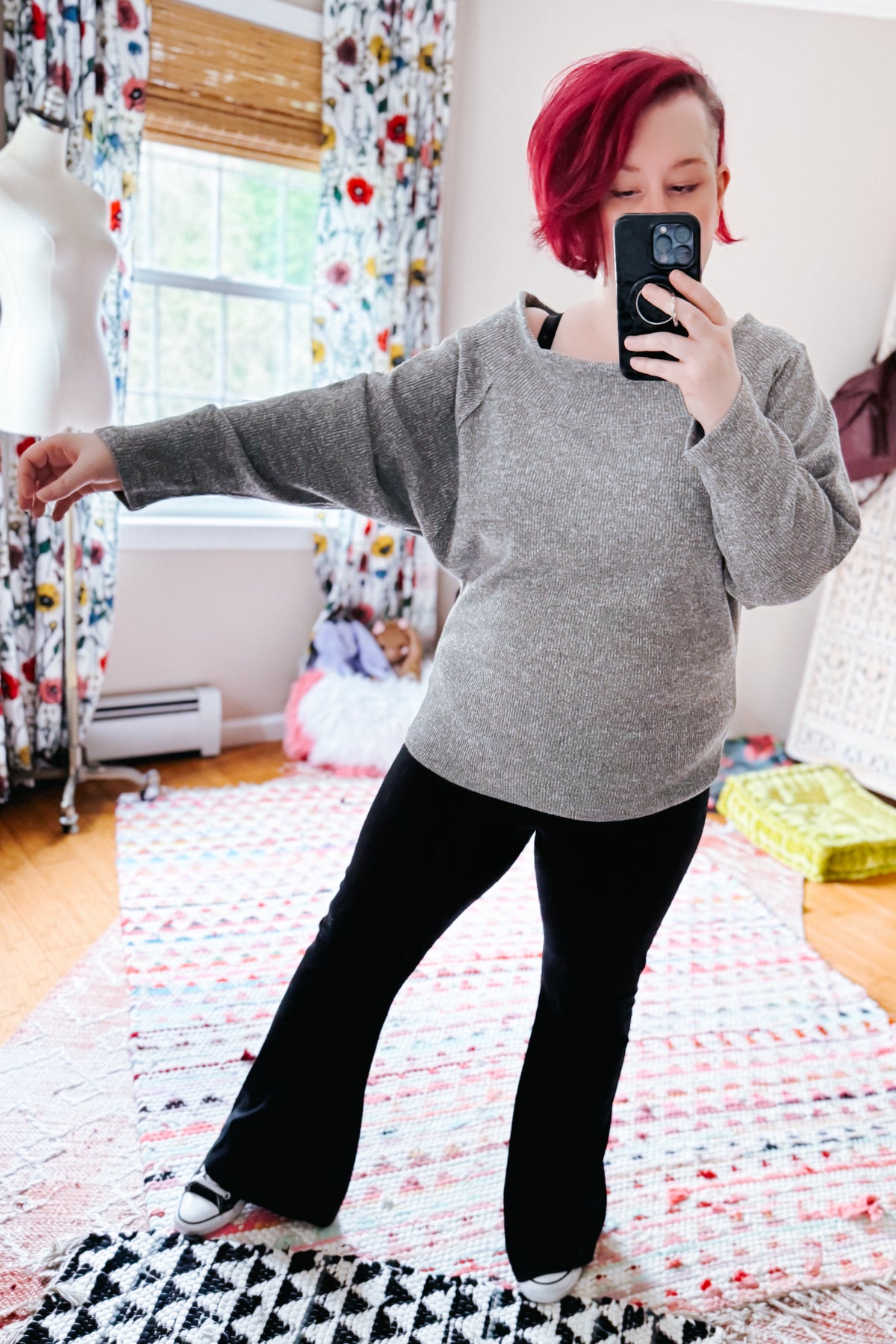 Woman taking a mirror selfie in a grey textured knit boat neck top with dolman sleeves and black pants indoors