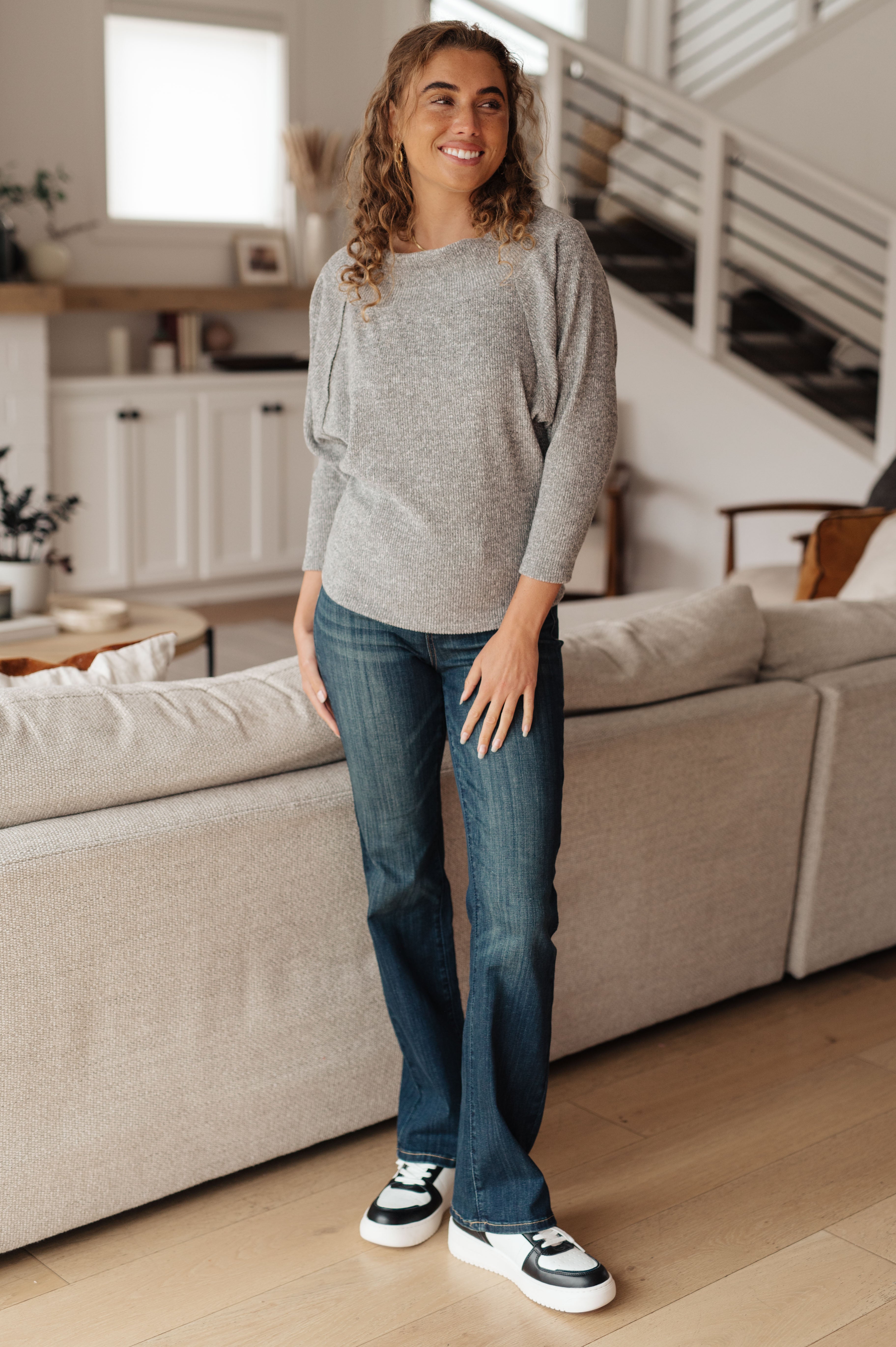 Woman wearing grey long sleeve top with relaxed fit and blue bootcut jeans standing indoors smiling