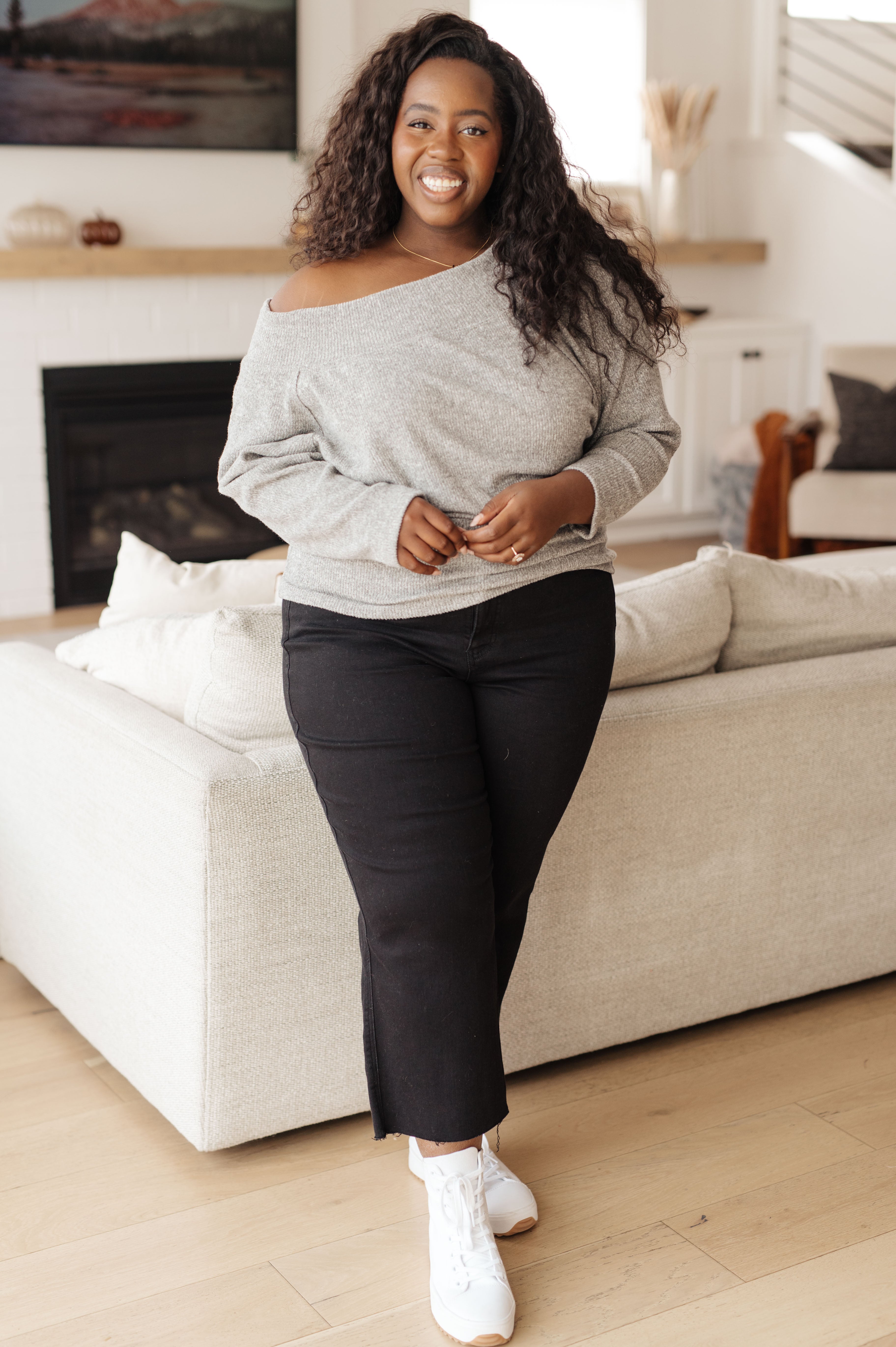 Smiling curvy woman wearing a grey off-shoulder long sleeve top and black pants in a stylish living room