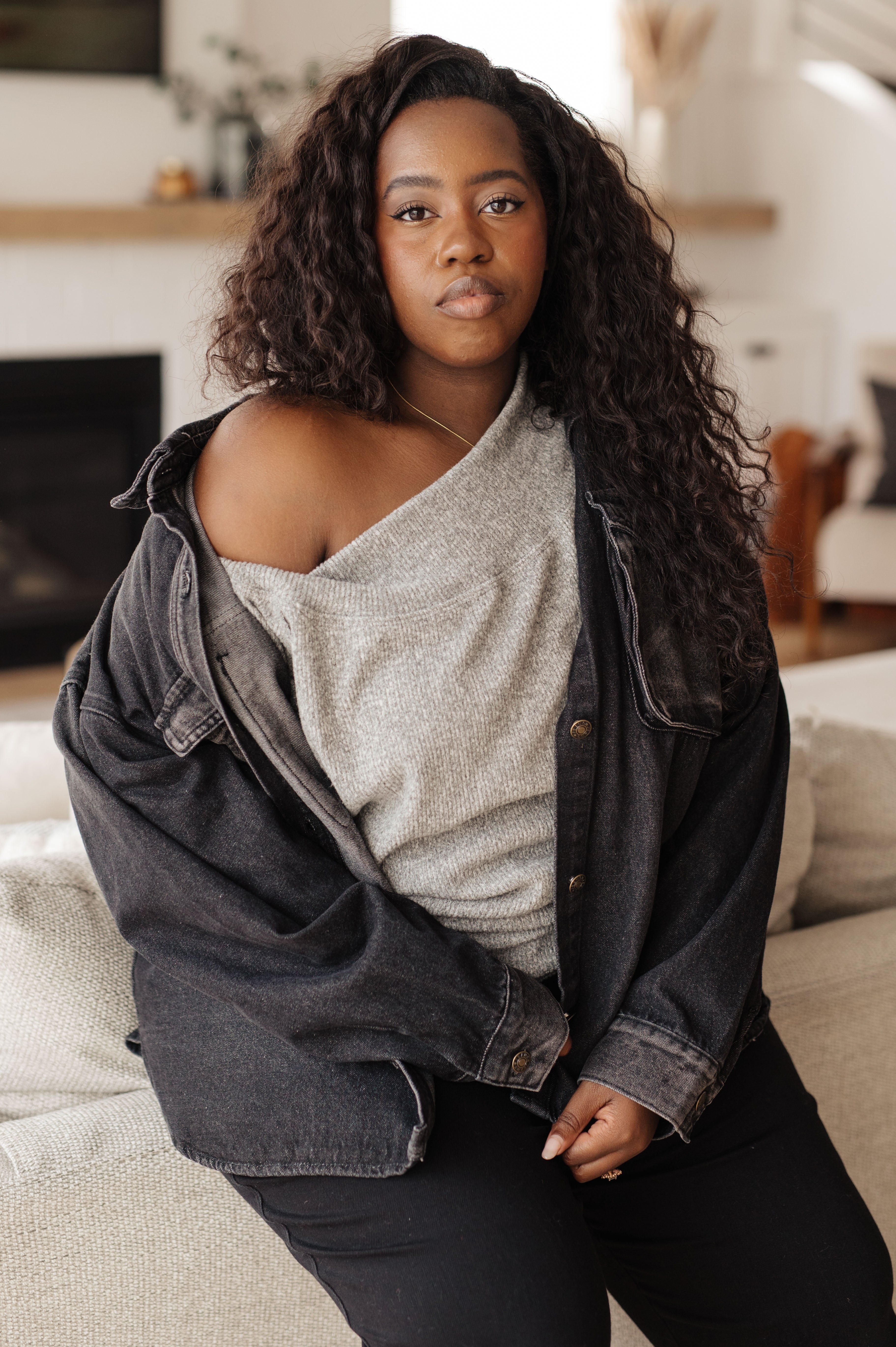 Confident woman wearing grey knit top with one shoulder exposed and oversized black jacket indoors in relaxed pose