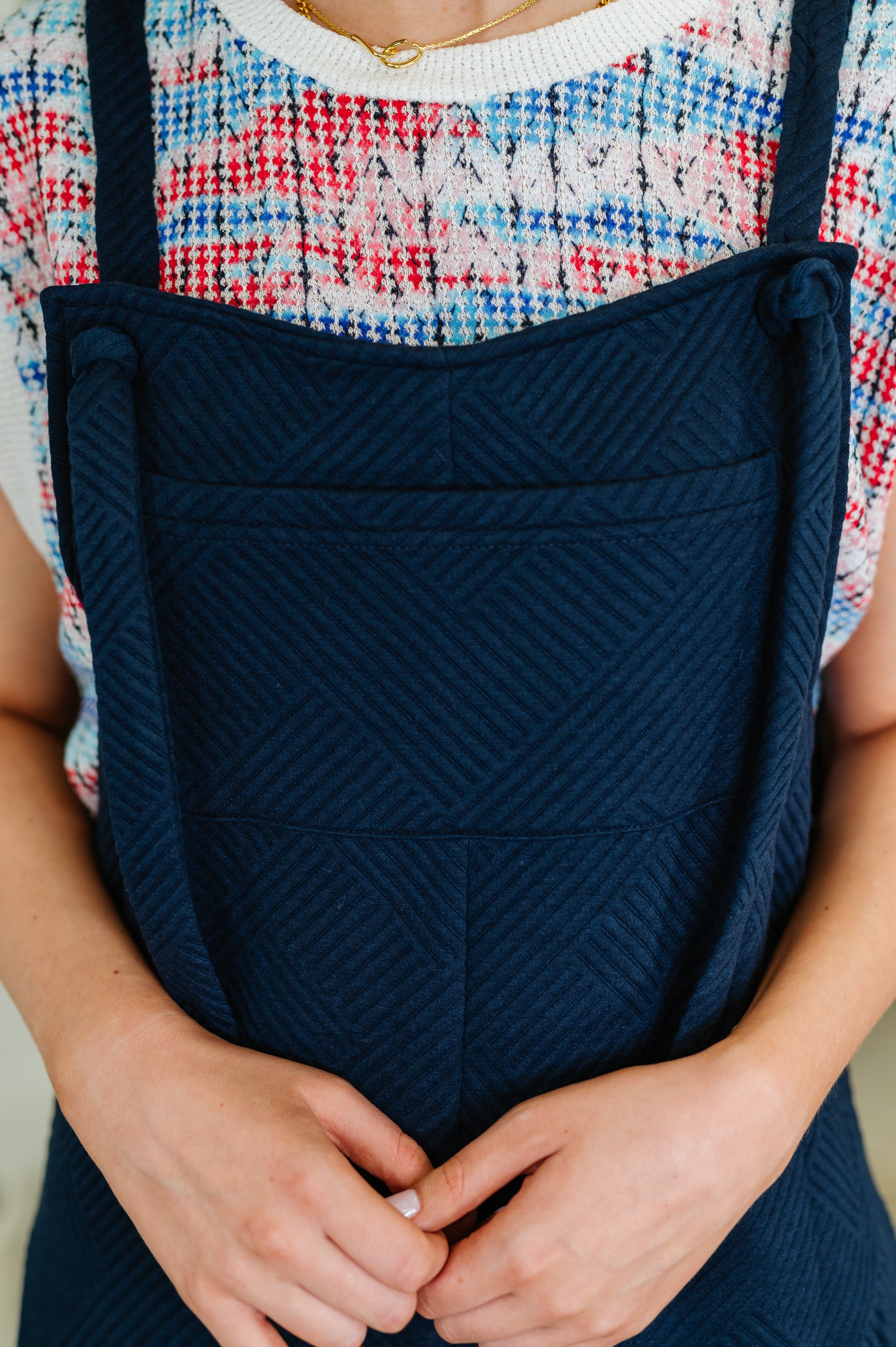 Close-up of navy textured fabric jumpsuit with adjustable straps worn over a colorful knit top with red and blue pattern