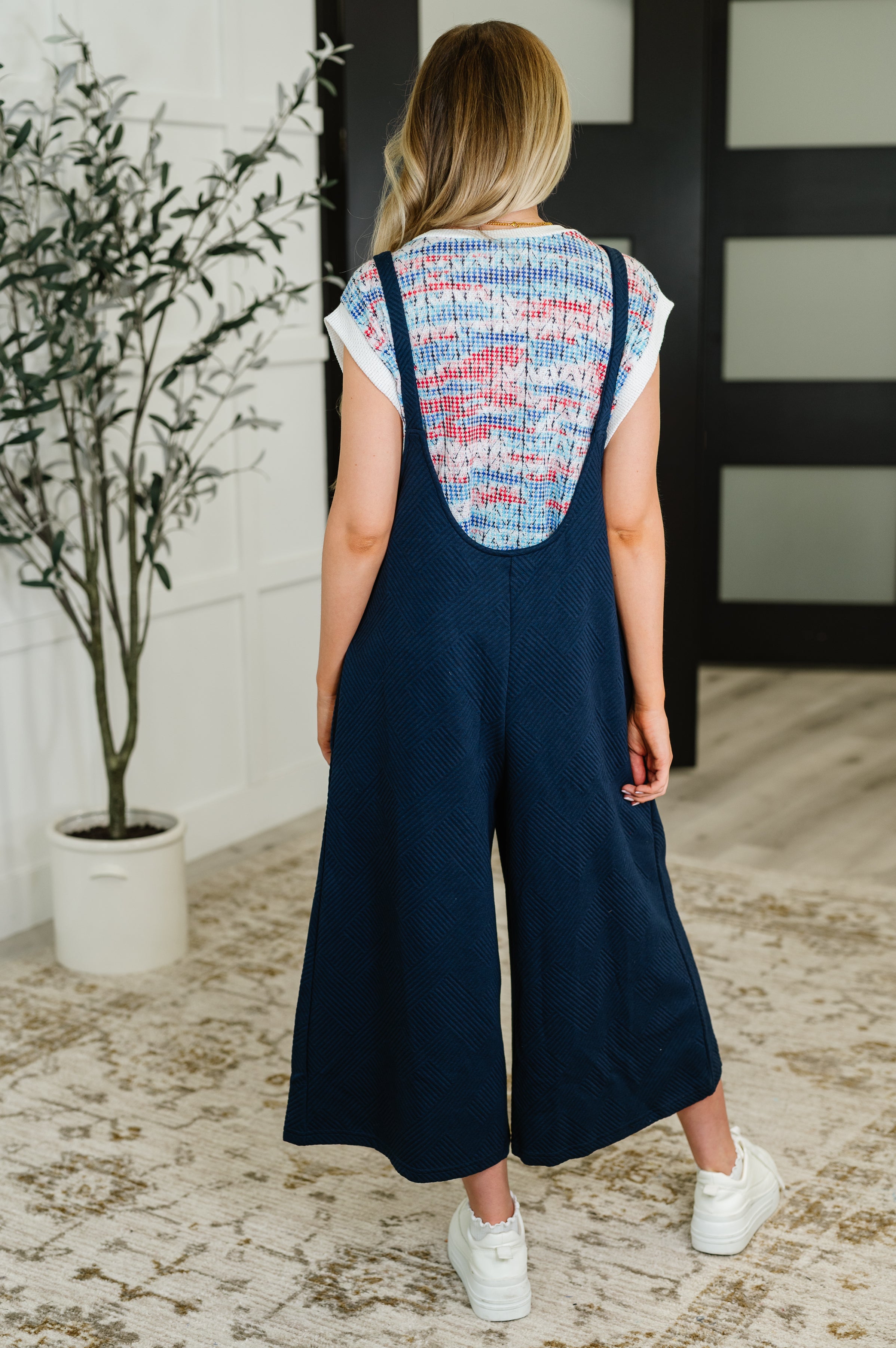 Woman wearing navy textured jumpsuit with adjustable straps and wide-leg pants standing indoors on rug