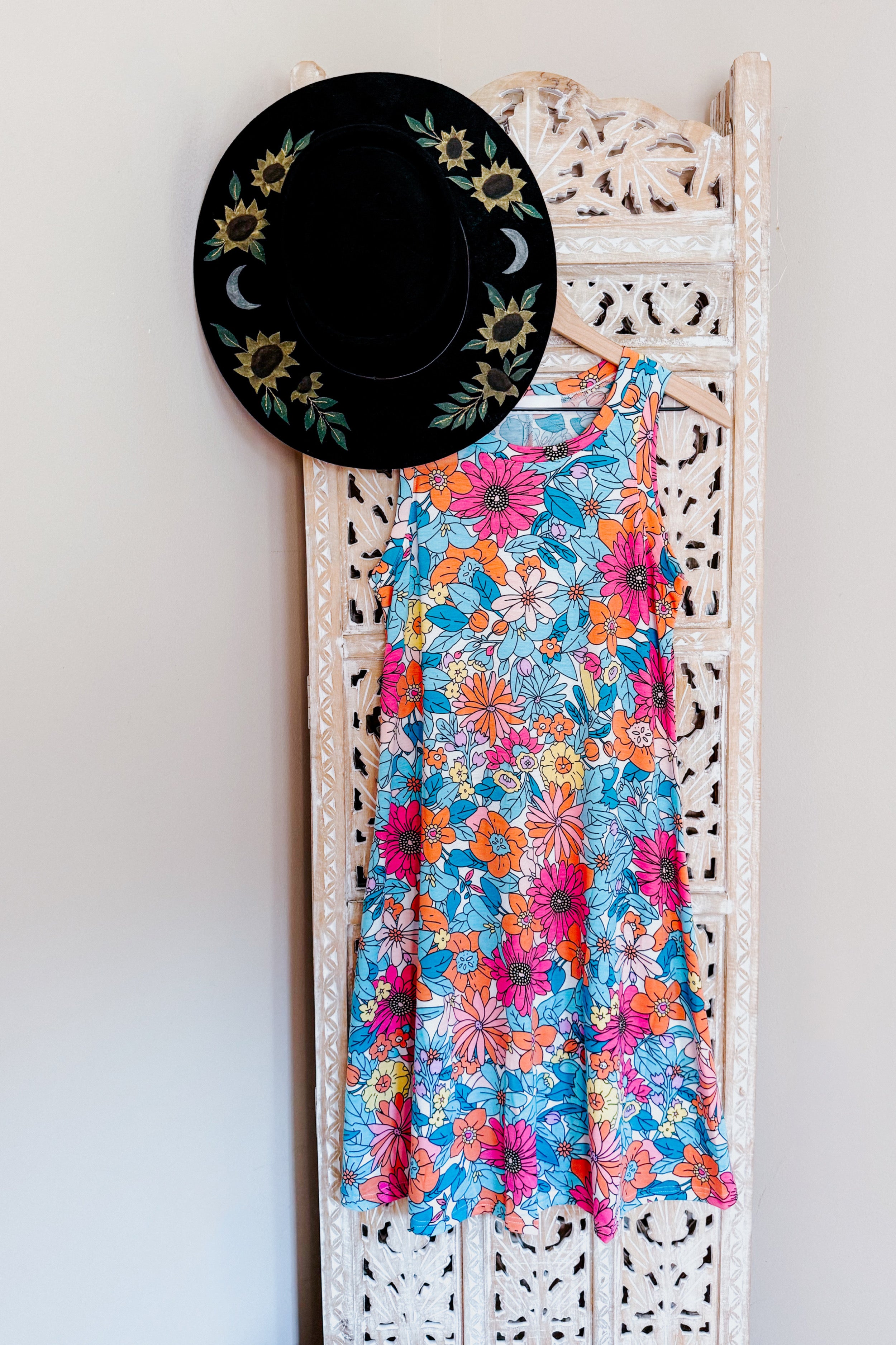 Blue sleeveless swing dress with bright floral print hanging on decorative carved wooden panel with black floral hat.