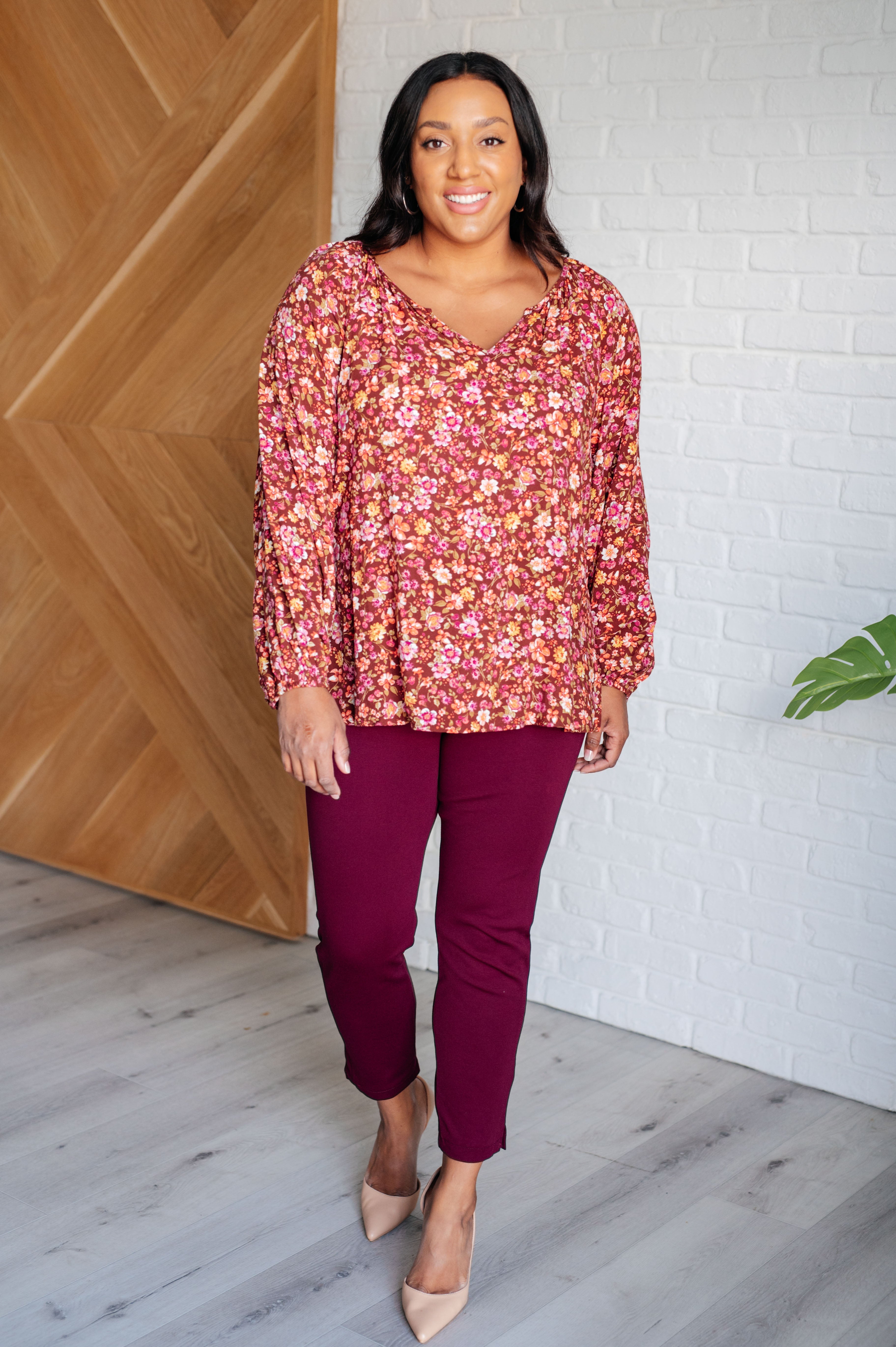 Curvy woman wearing a rust floral blouse with long sleeves paired with burgundy pants and beige heels indoors