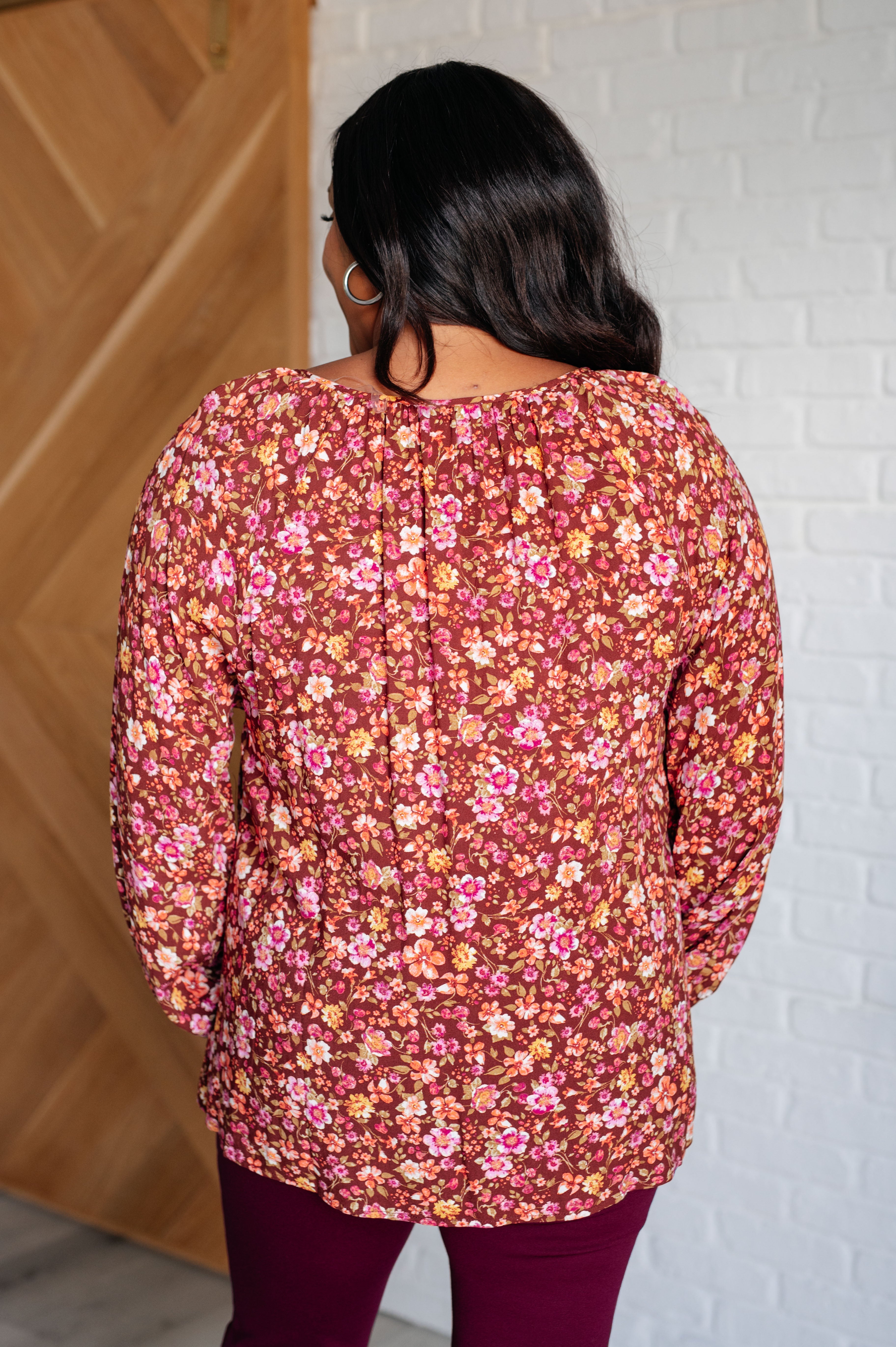 Woman wearing a rust floral blouse with pink and orange flowers standing indoors with back facing camera