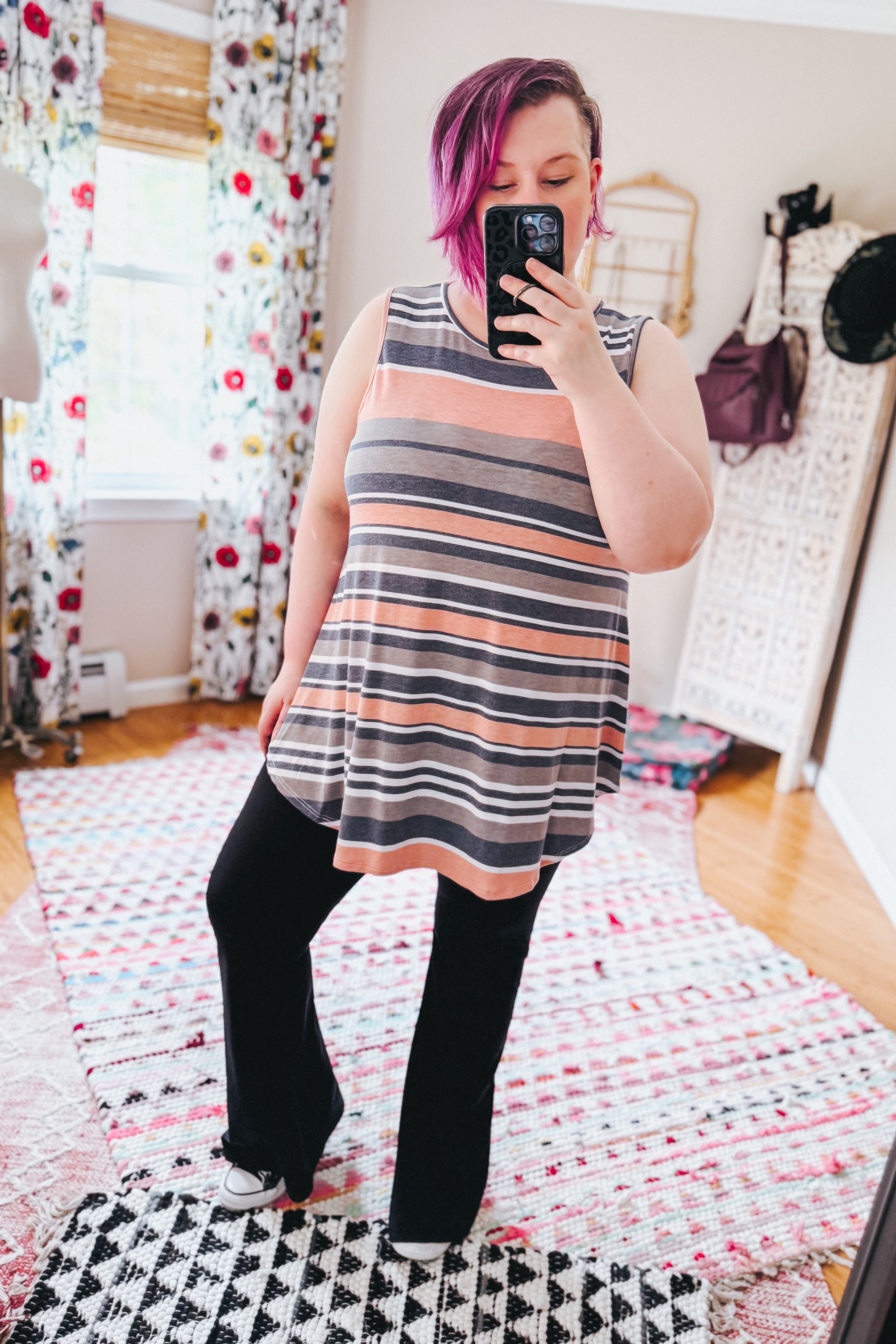 Woman with pink hair wearing a sleeveless striped top and black pants taking a mirror selfie in a decorated room