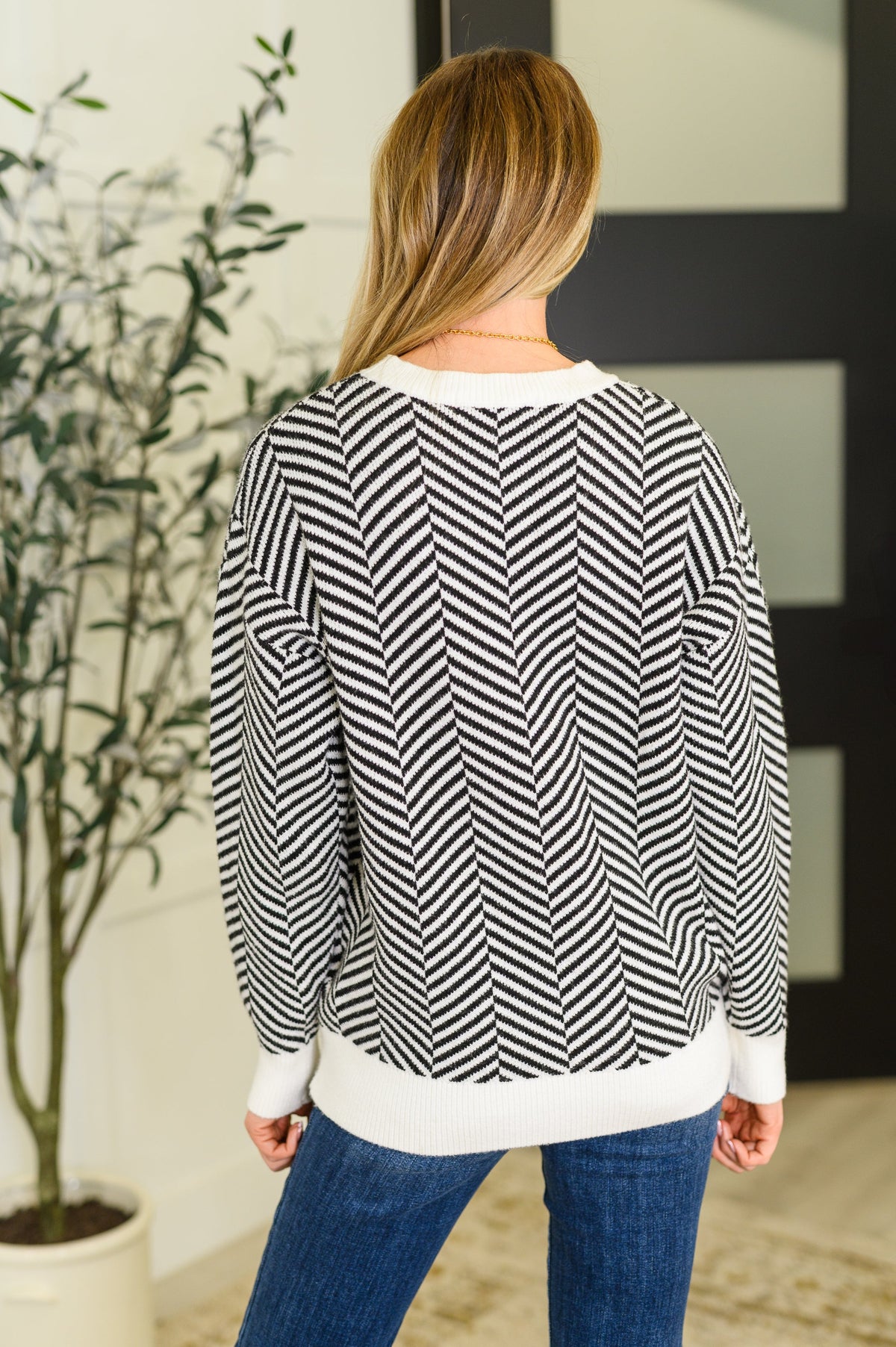 Woman viewed from behind wearing a black and white chevron knit sweater with ribbed cuffs and hem and blue jeans indoors