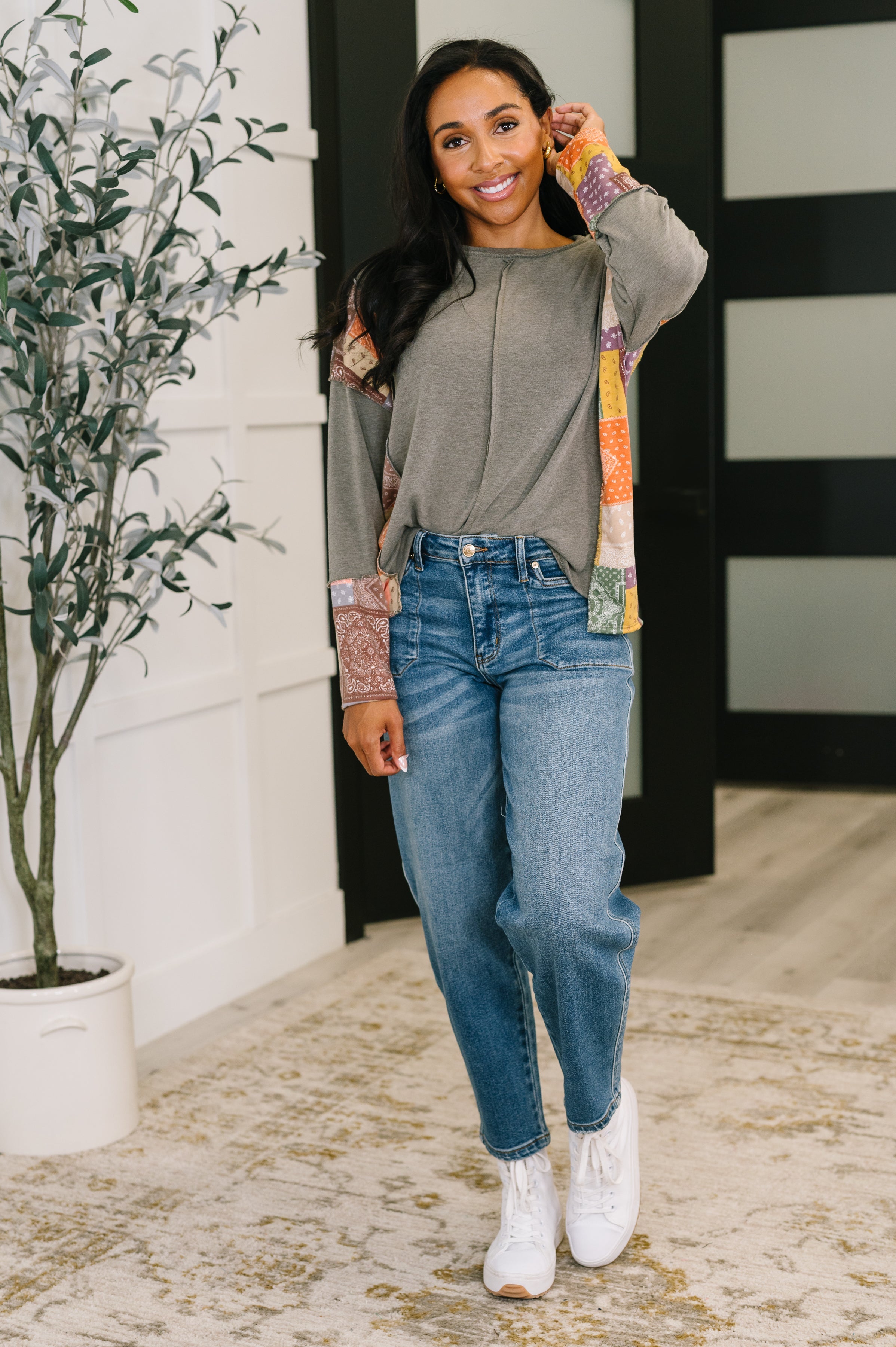 Smiling woman wearing an olive color block relaxed long sleeve with patchwork sleeves and blue jeans indoors