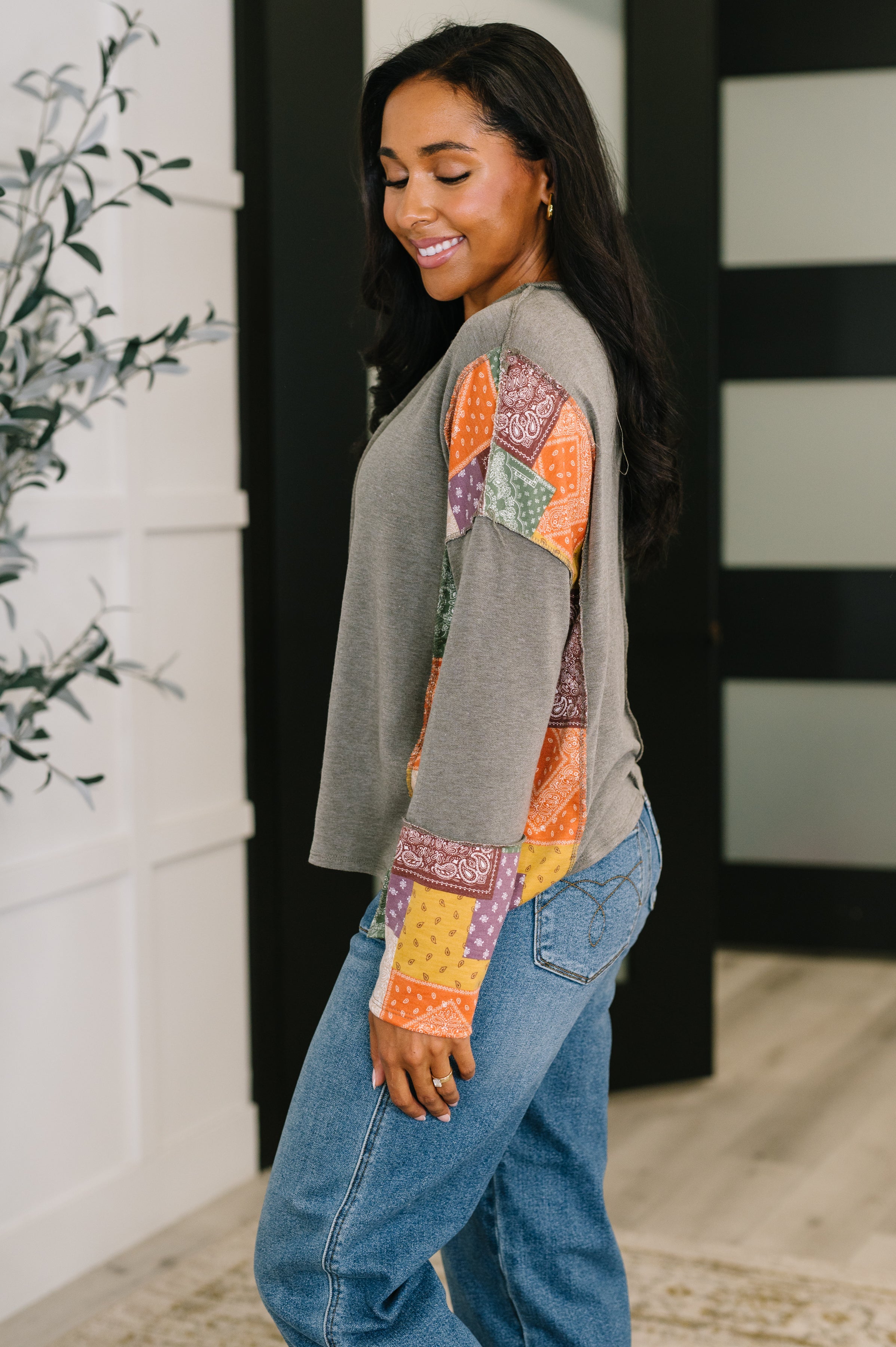 Woman wearing an olive toned relaxed long sleeve with colorful patchwork sleeves paired with blue jeans indoors