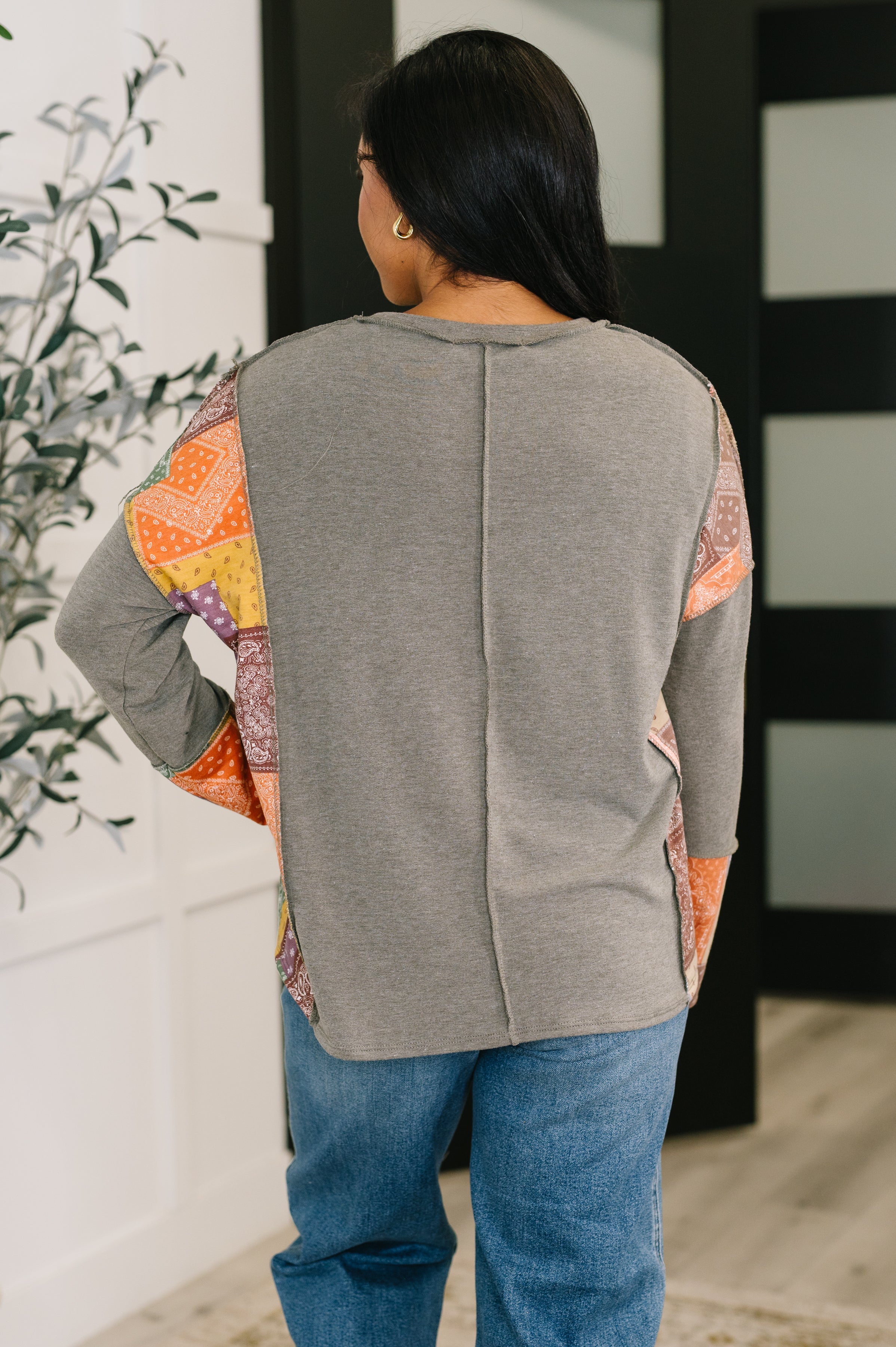 Woman wearing an olive long sleeve top with colorful patchwork sleeves paired with relaxed blue jeans indoors