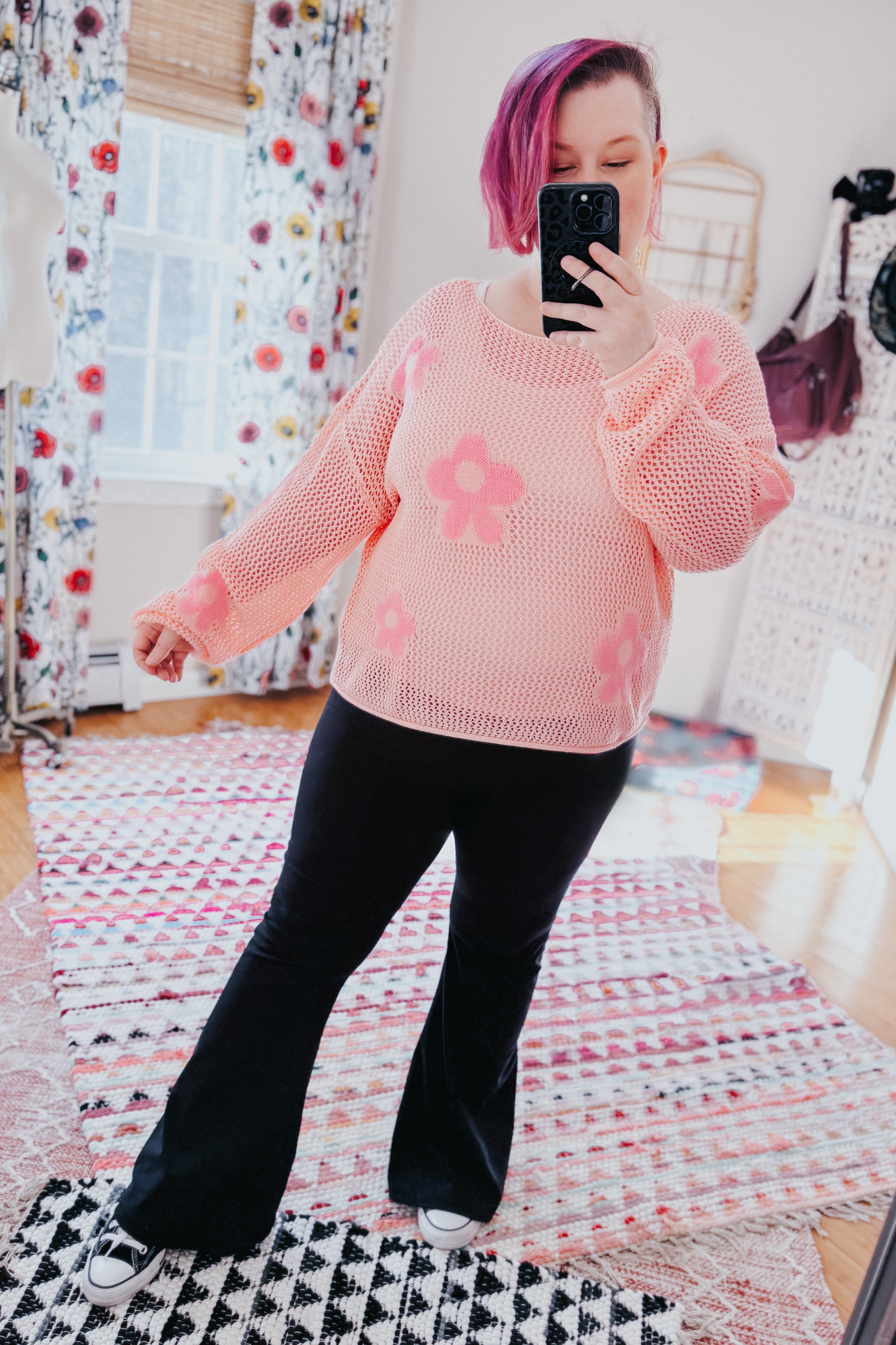Woman wearing a blush pink mesh sweater with floral patterns paired with black flared pants indoors taking a mirror selfie