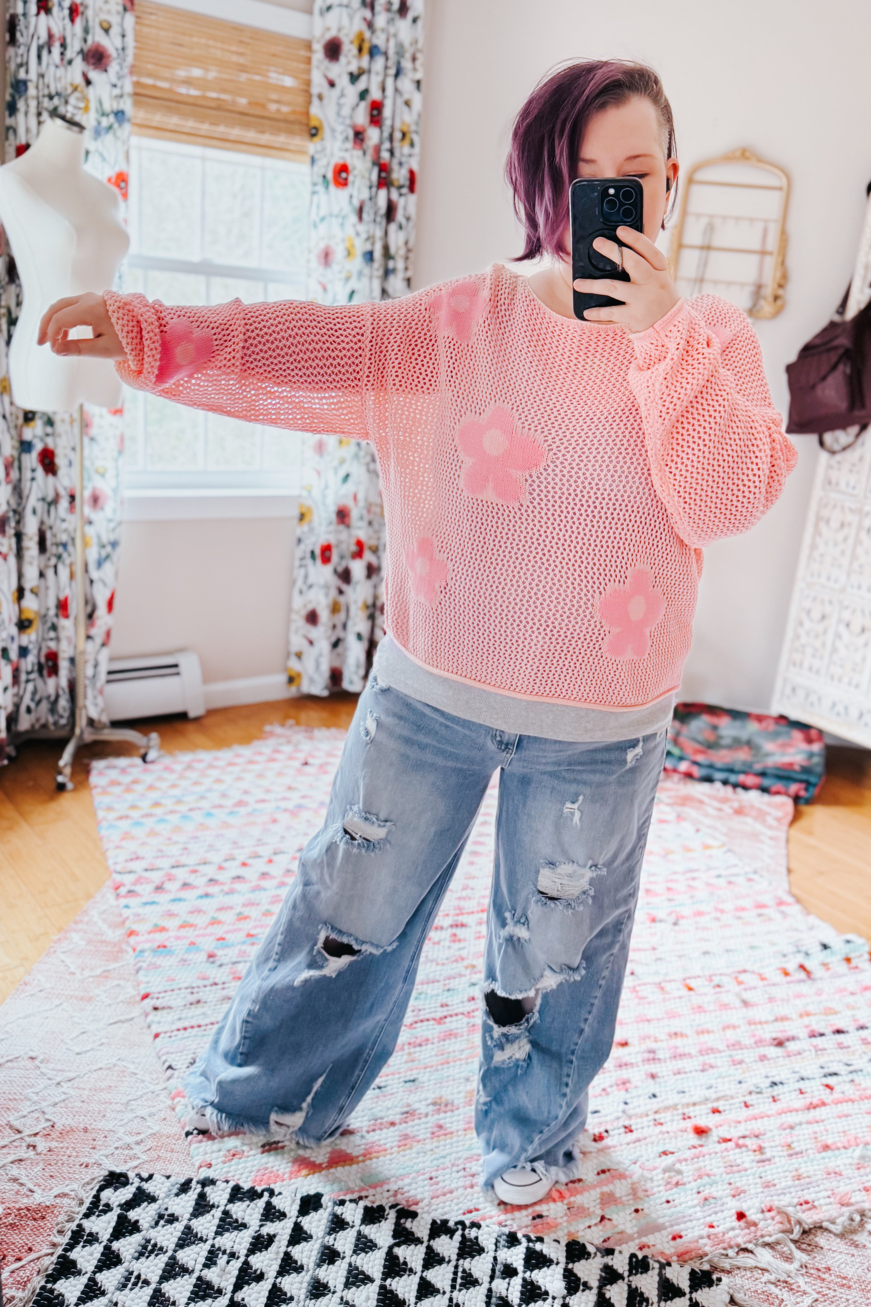 Woman wearing a blush pink floral mesh sweater top with loose ripped denim jeans indoors taking a mirror selfie