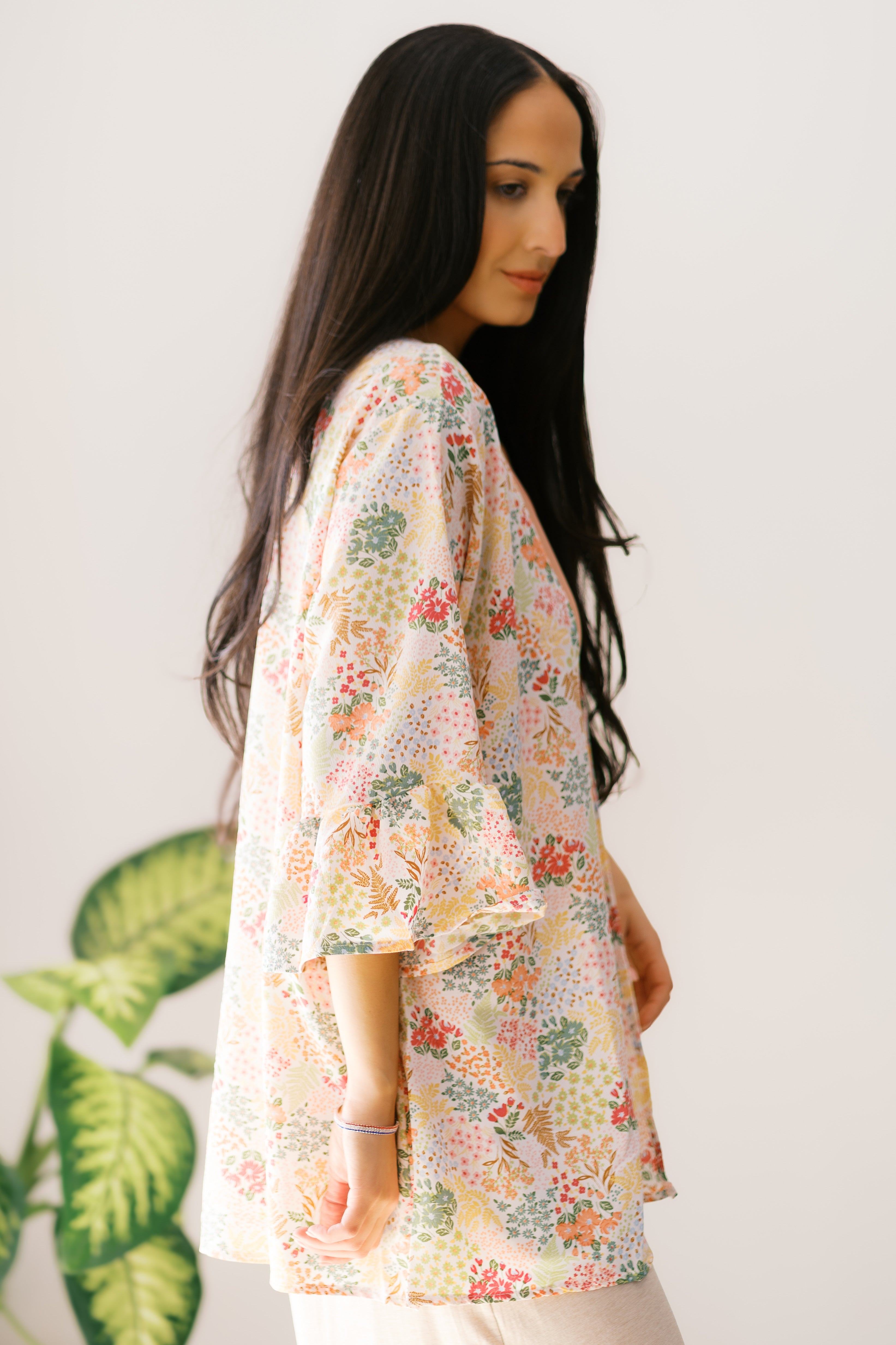 Woman wearing an ivory floral lightweight cardigan with ruffled sleeves standing next to green foliage indoors