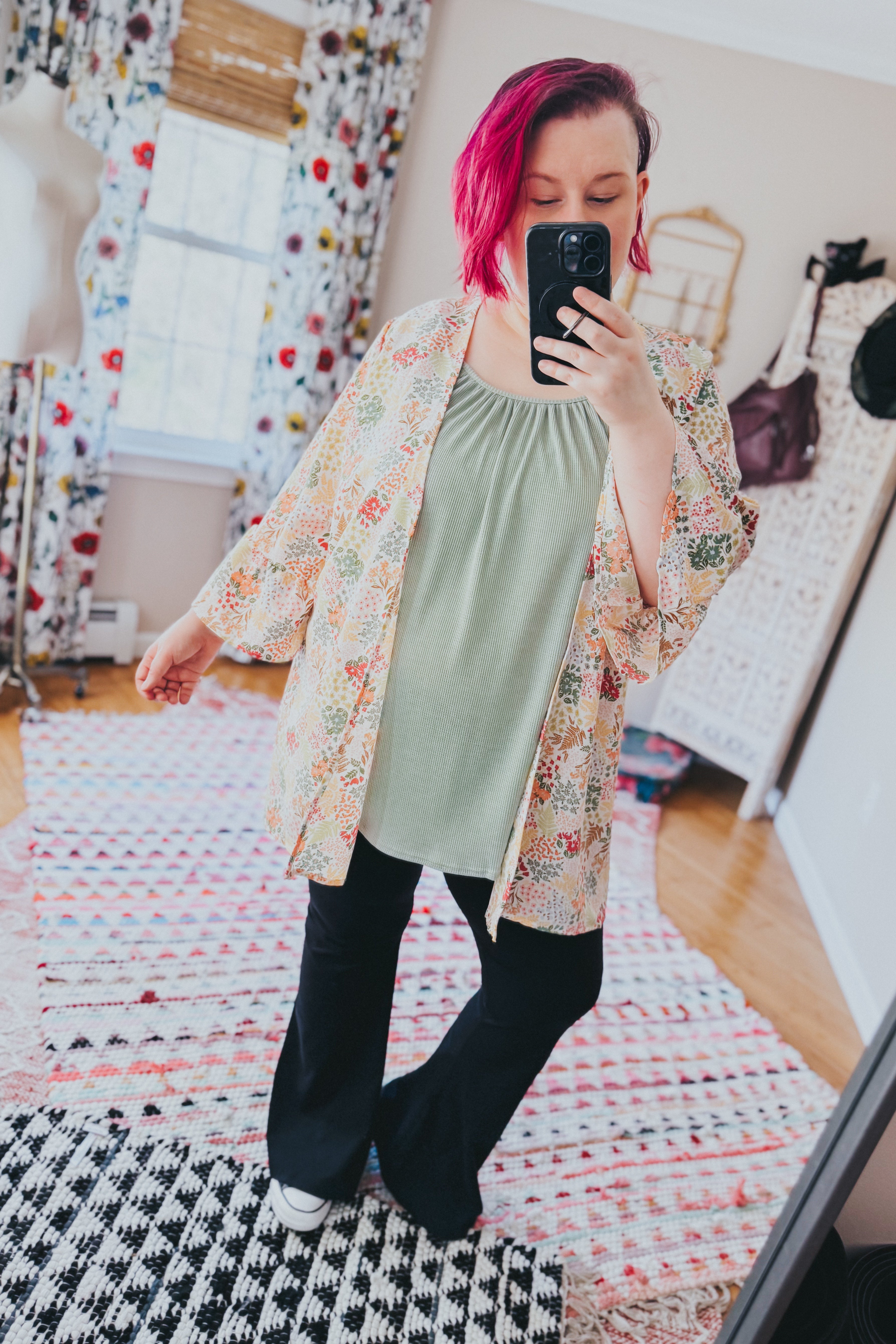 Woman with short magenta hair wearing an ivory floral light cardigan over a sage green top and black pants indoors taking a selfie
