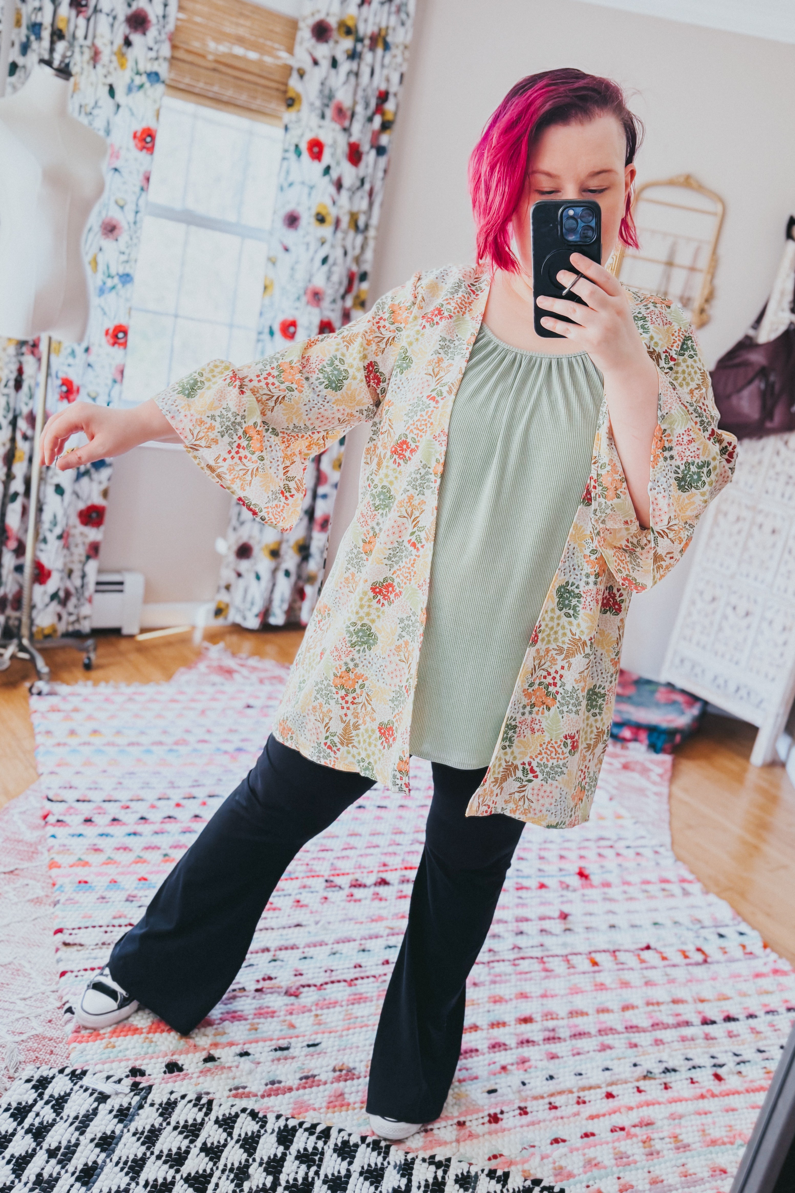 Woman with bright pink hair wearing an ivory floral light cardigan over a sage top and black flared pants indoors
