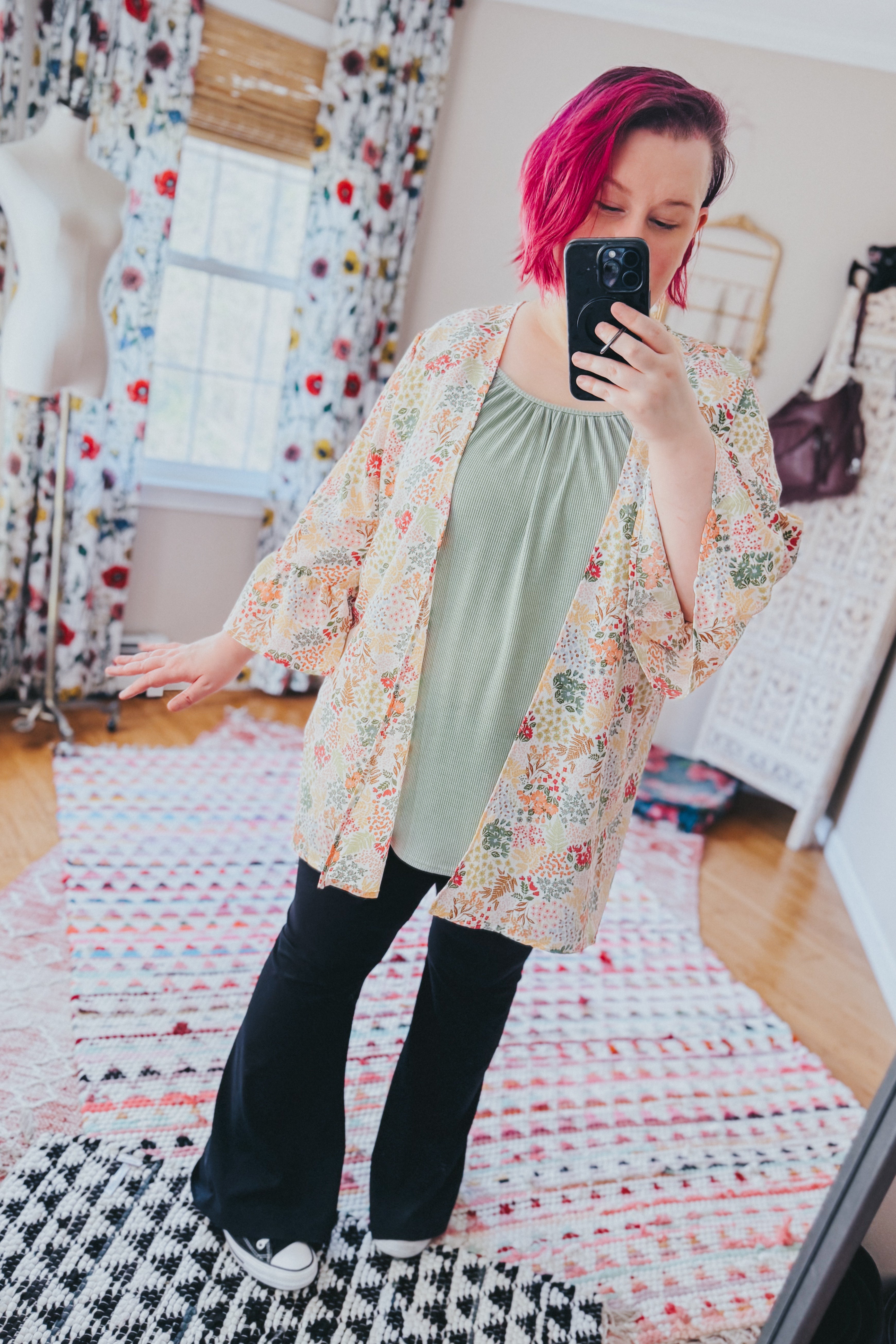Woman with short pink hair wearing an ivory floral light cardigan over sage top and black pants indoors taking a selfie