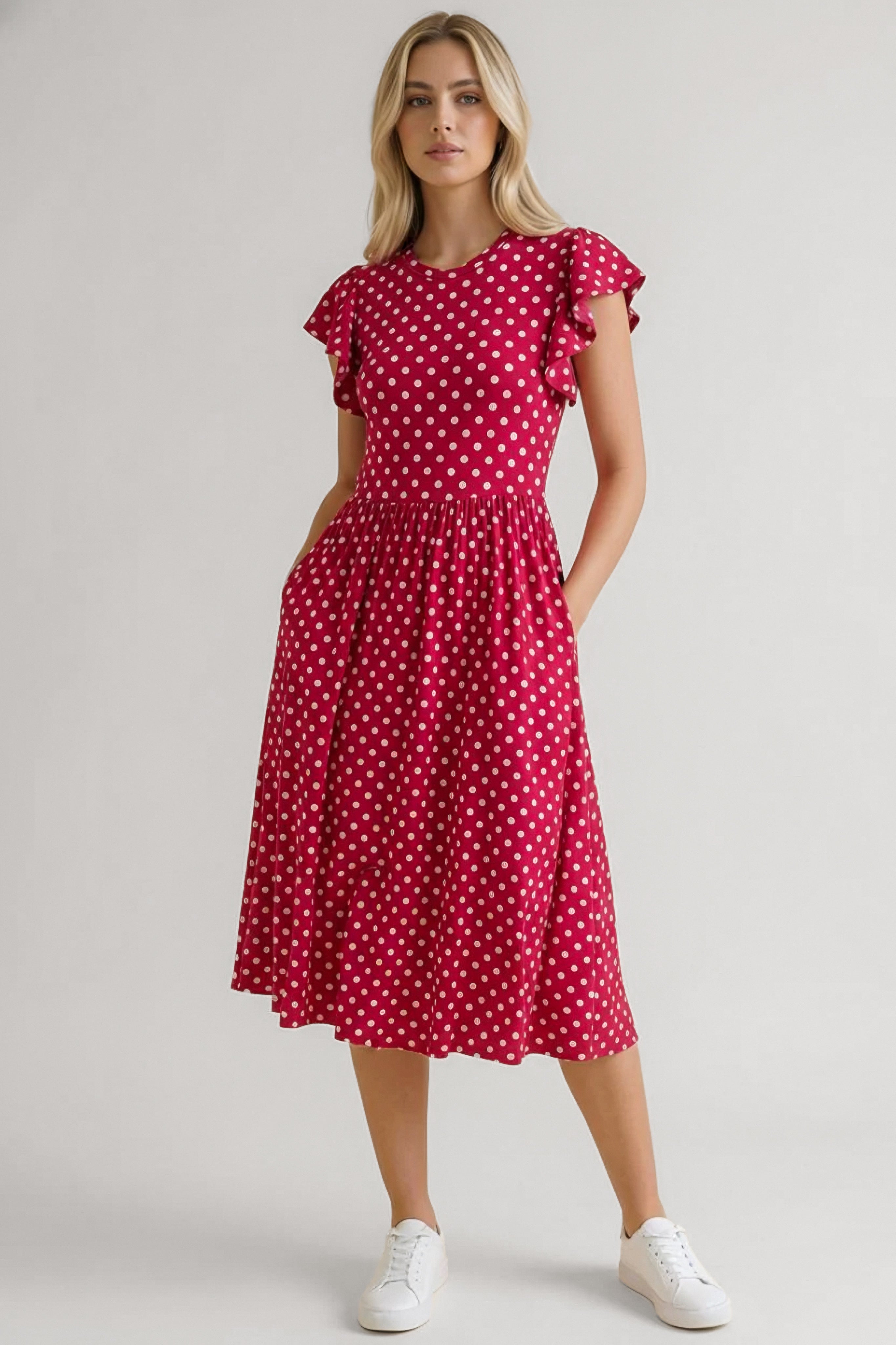 Woman wearing a red polka dot midi dress with flutter sleeves and gathered waist paired with white sneakers standing confidently