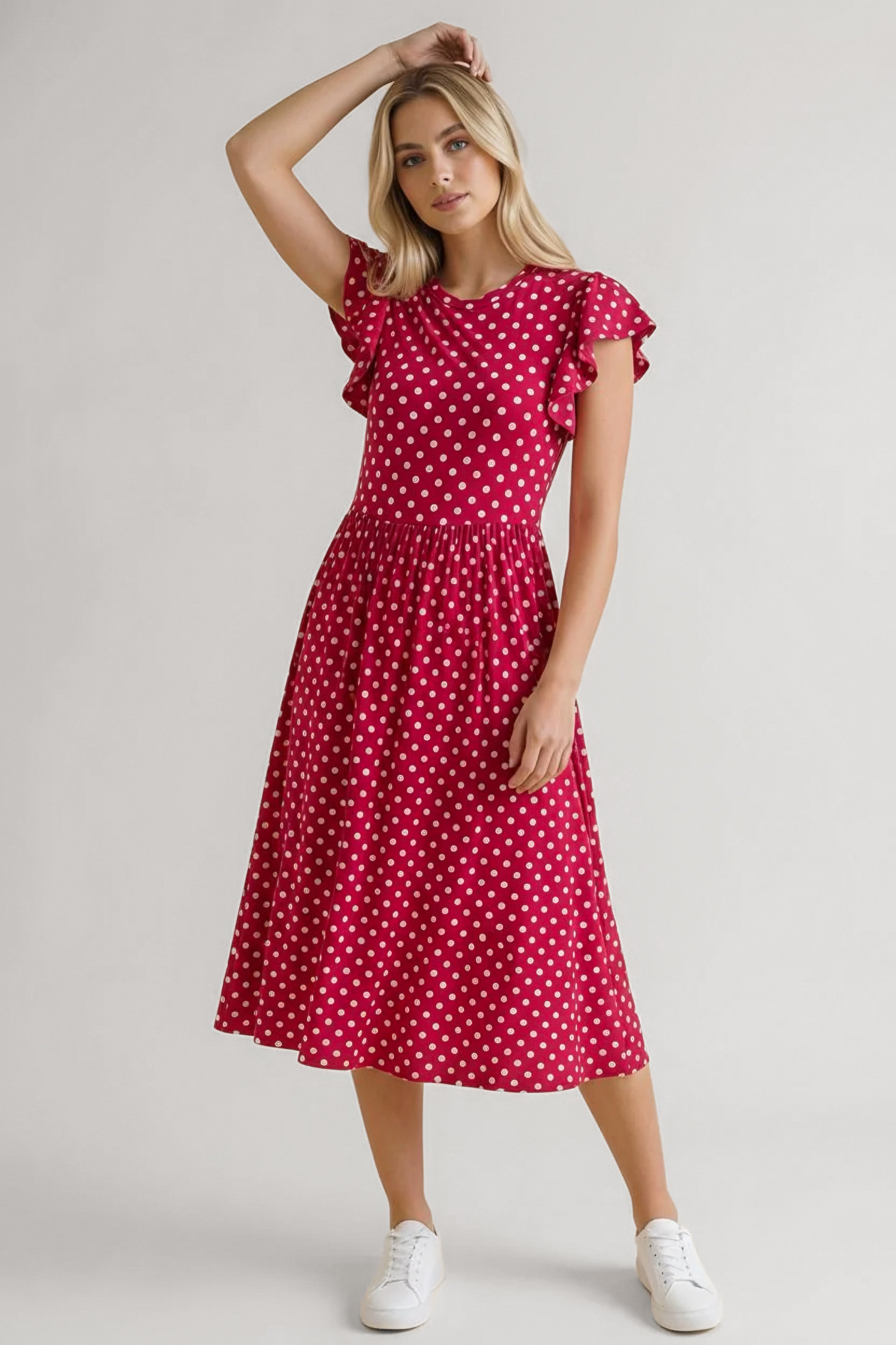 Woman wearing red polka dot midi dress with flutter sleeves and gathered waist paired with white sneakers, posing confidently