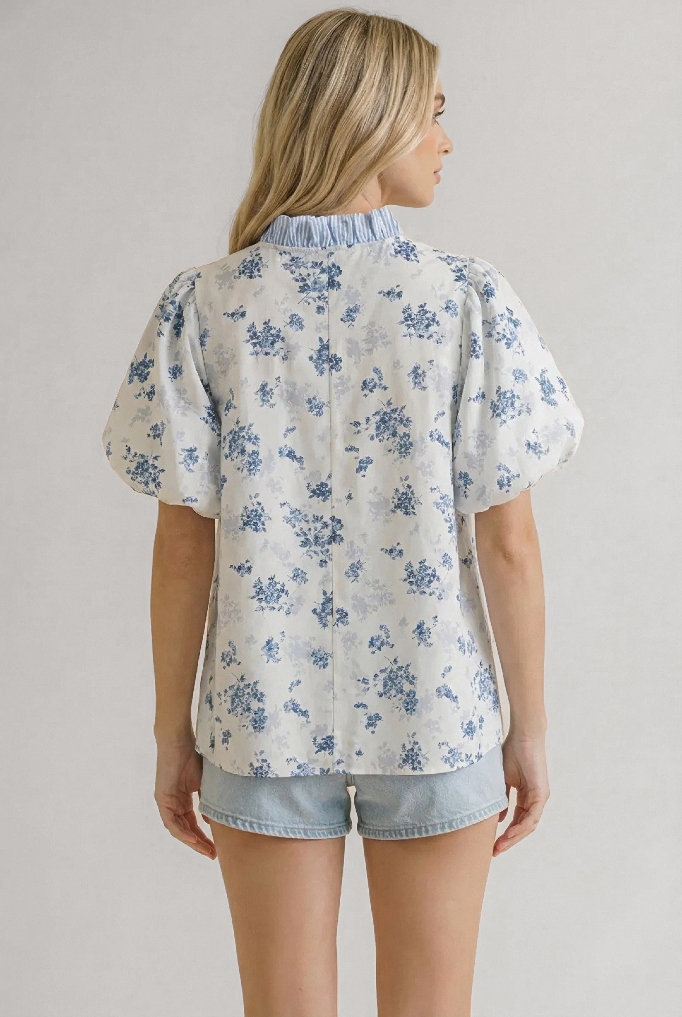 Woman wearing white blouse with blue floral print and puffed sleeves paired with light denim shorts viewed from back
