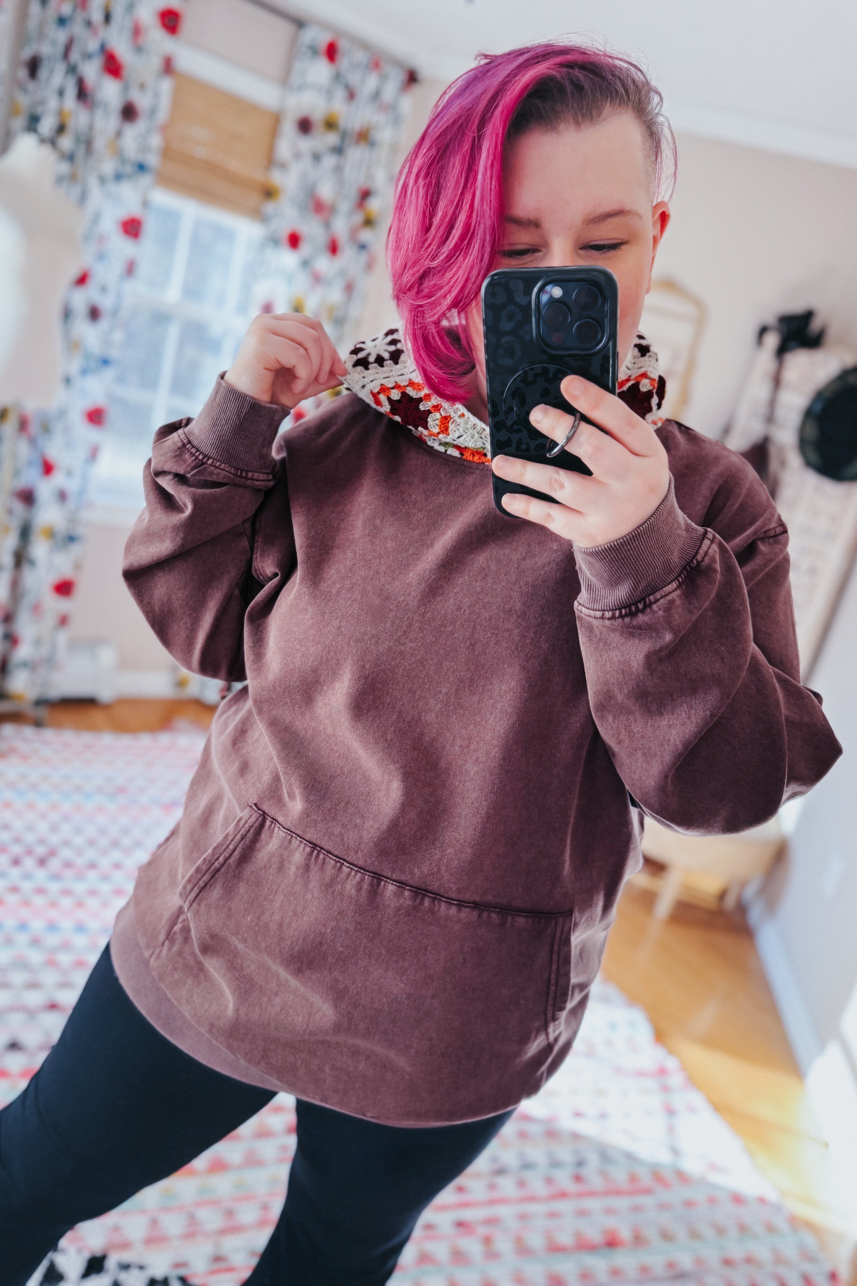 Woman with pink hair wearing a cozy acid wash hoodie with crochet hood taking a mirror selfie indoors