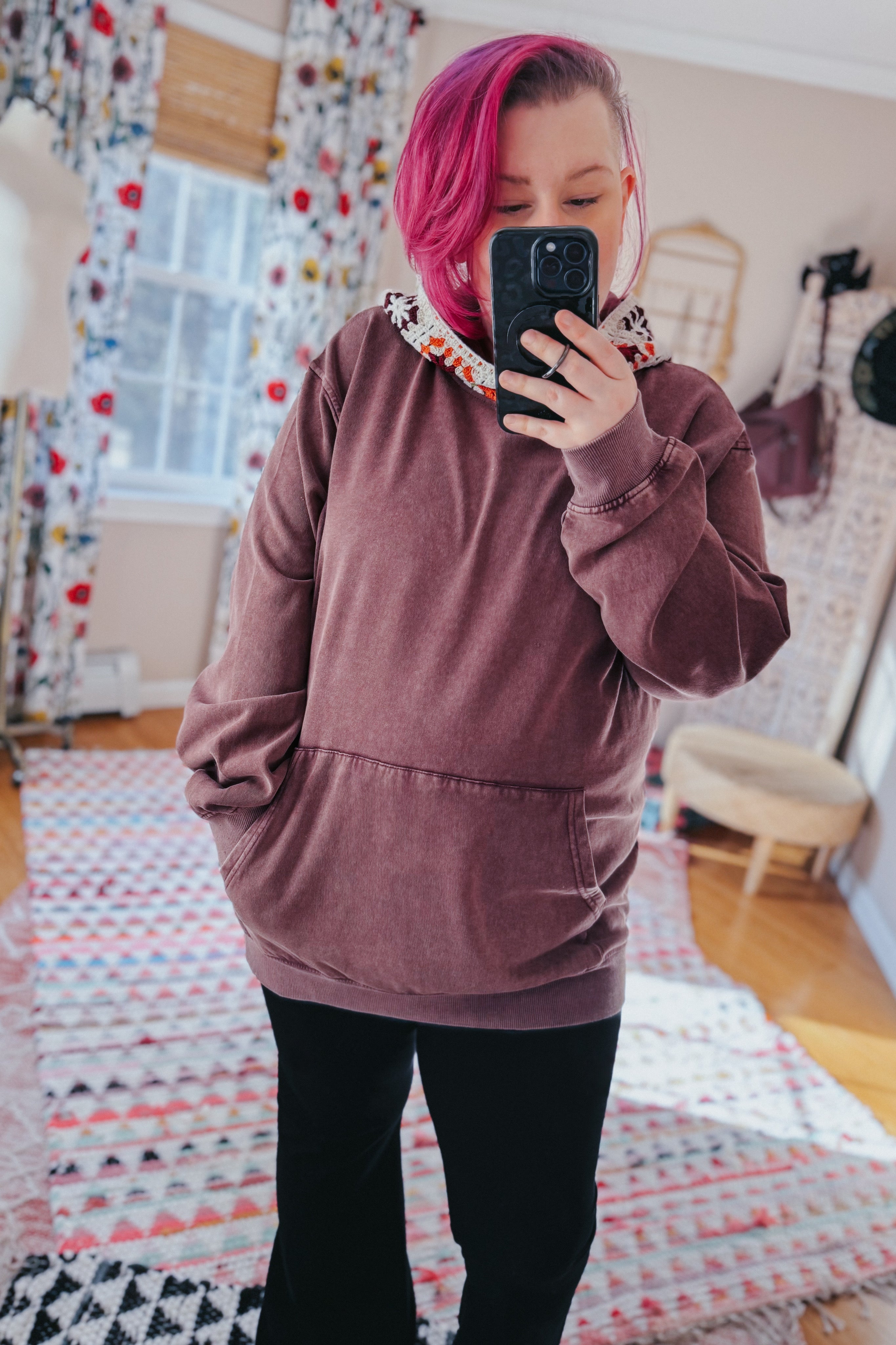 Woman with pink hair wearing a plush acid wash hoodie with crochet hood detail taking a mirror selfie indoors