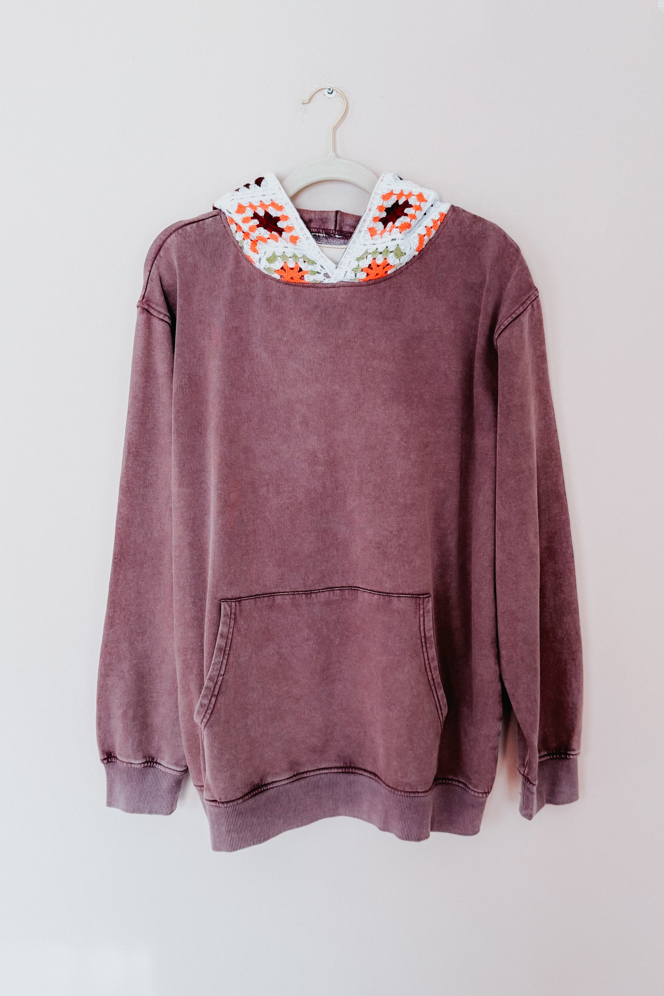 Purple acid wash hoodie with front pocket and white crochet hood featuring orange and green accents hanging on white wall