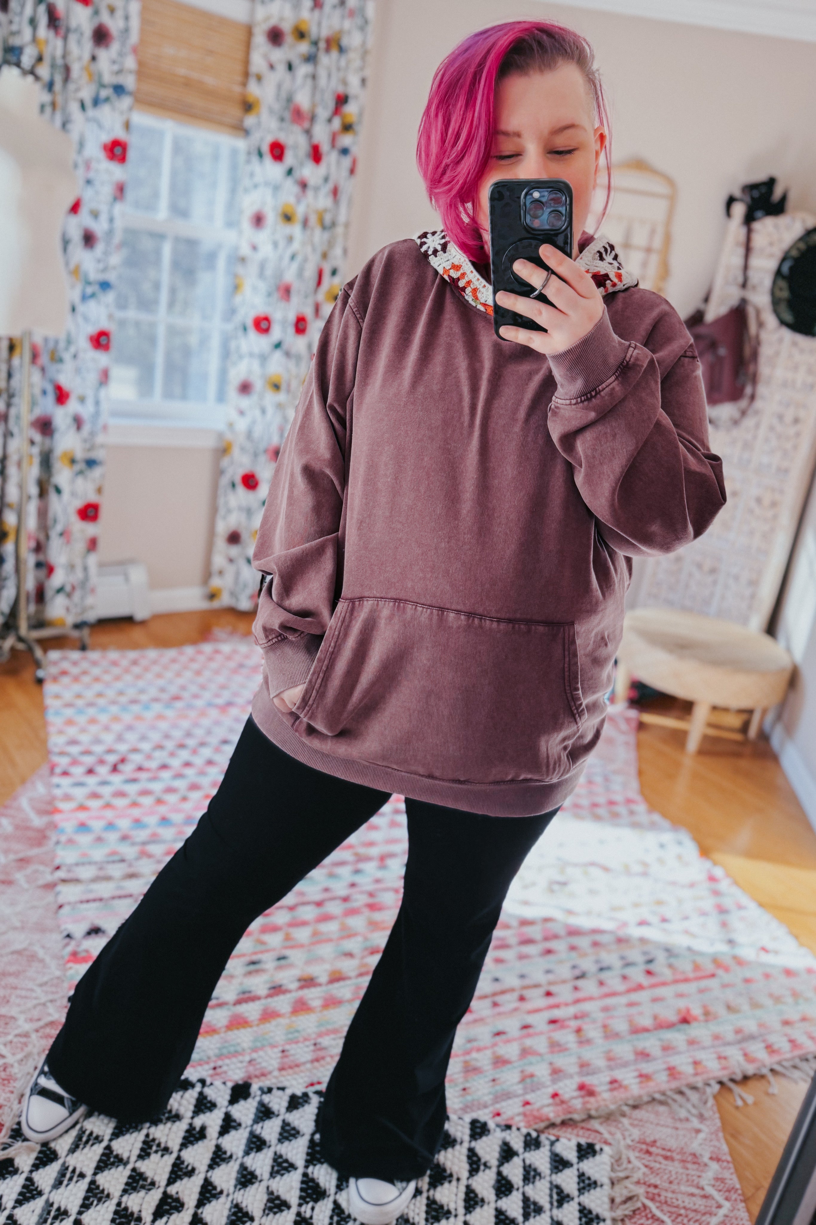 Woman with pink hair wearing a purple acid wash hooded hoodie with floral trim paired with black flared pants indoors