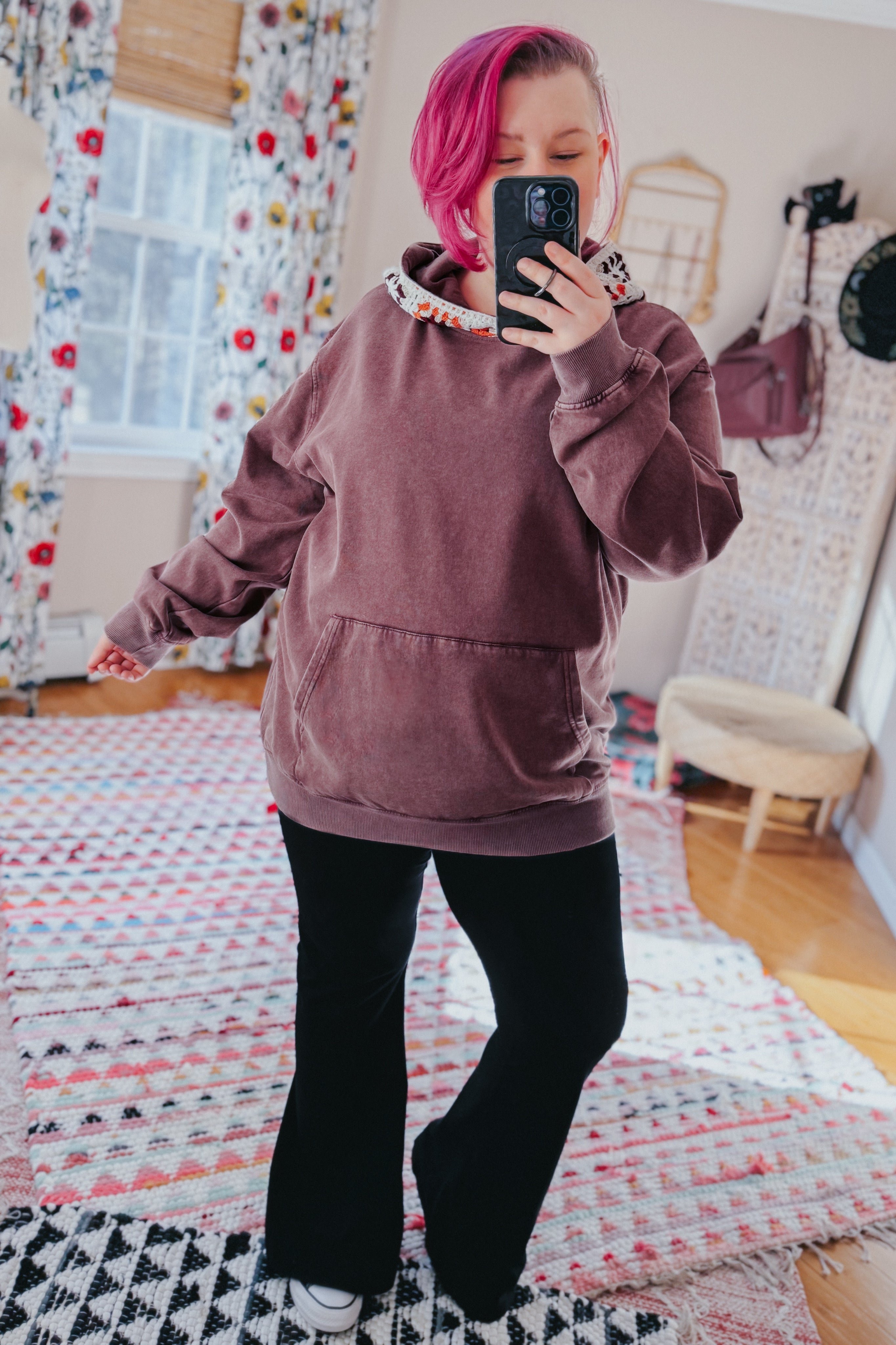 Woman with pink hair wearing a plush acid wash hoodie with floral hood lining and black pants taking a mirror selfie indoors