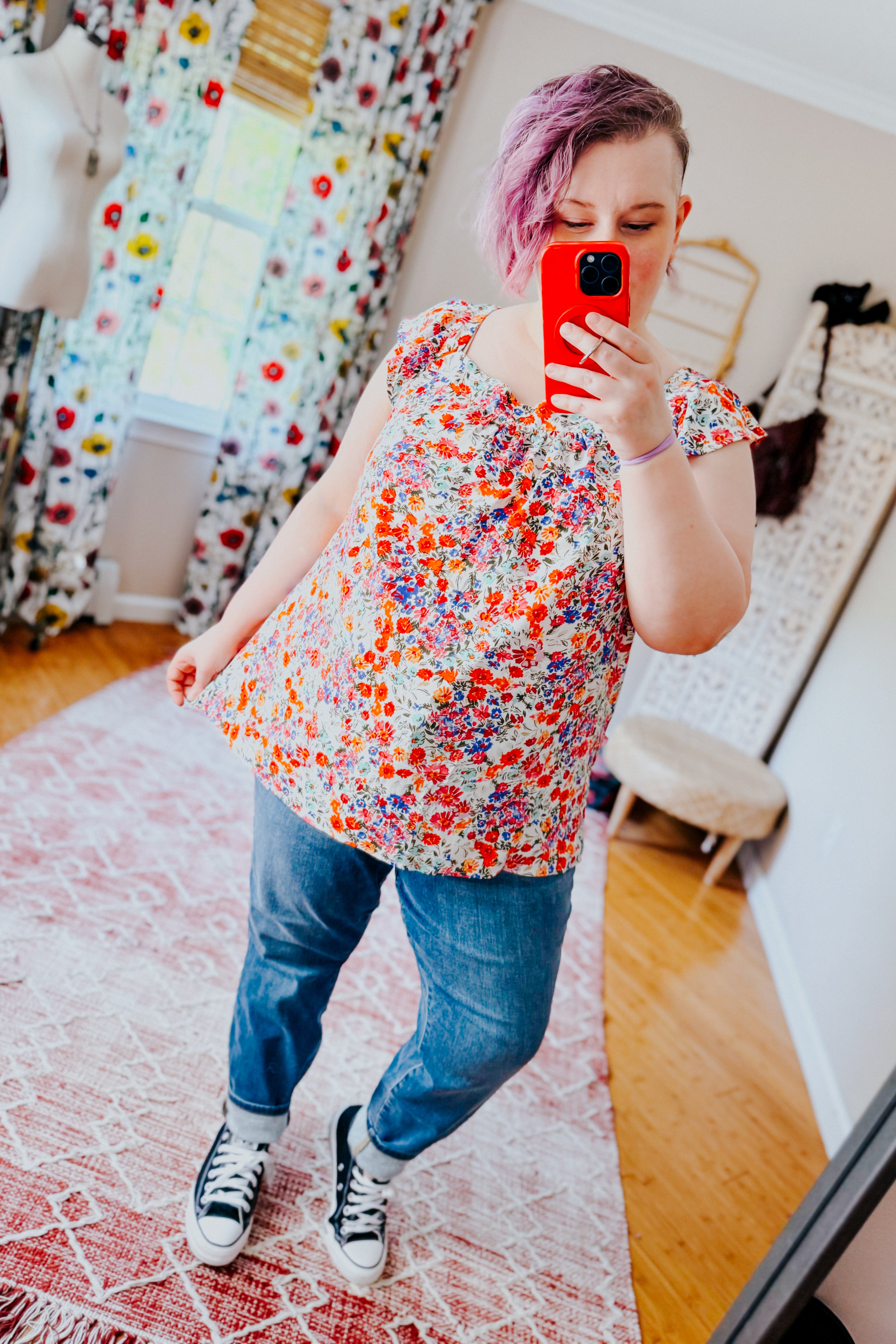 Woman with short pink hair wearing a red floral square neck top, blue jeans, and black sneakers taking a mirror selfie indoors
