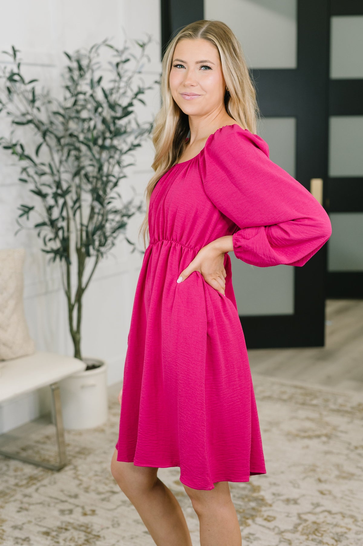 Woman modeling a pink square neck dress with long raglan sleeves and a gathered waist in a bright modern home interior