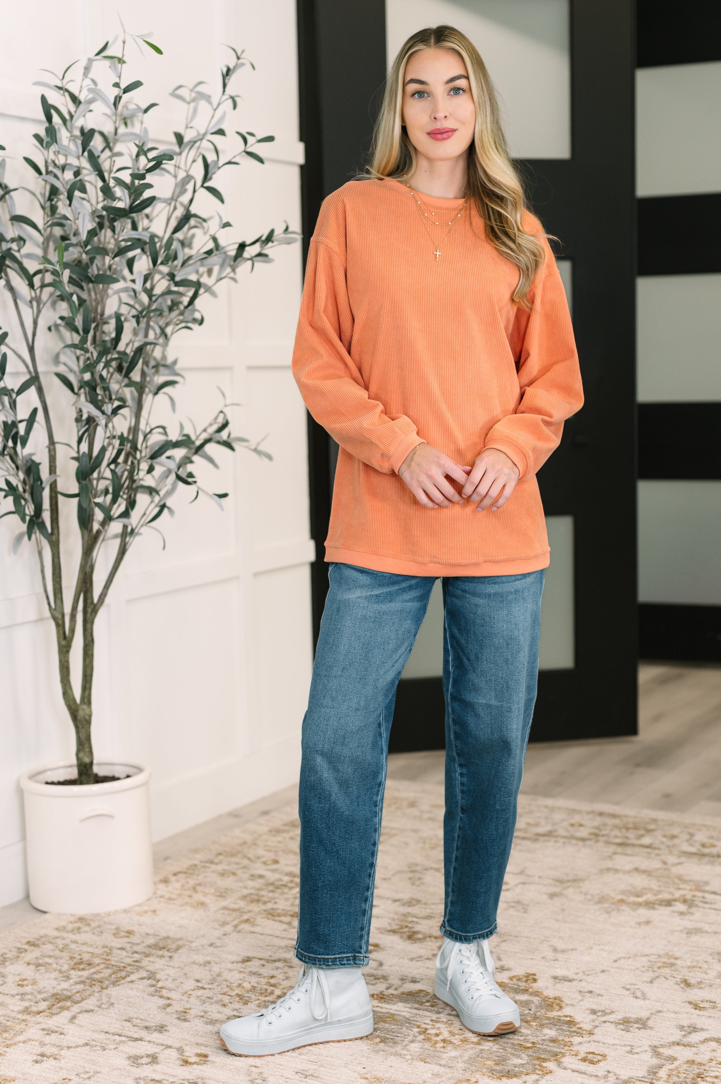 Woman wearing an oversized orange corded sweatshirt paired with blue jeans and white sneakers in stylish indoor setting