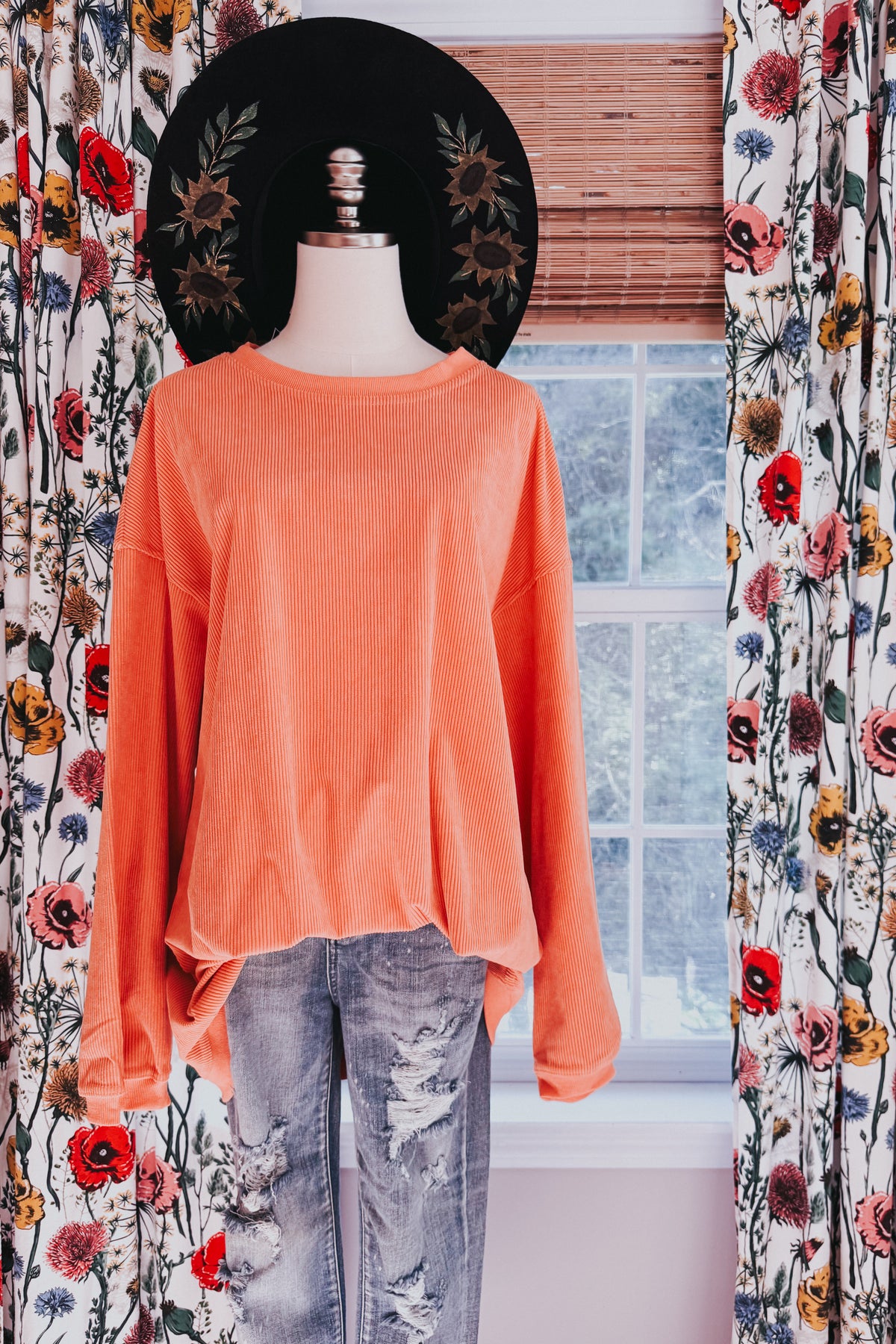 Orange oversized corded sweatshirt with a ribbed round neckline and drop shoulders styled with distressed jeans on a mannequin by a window