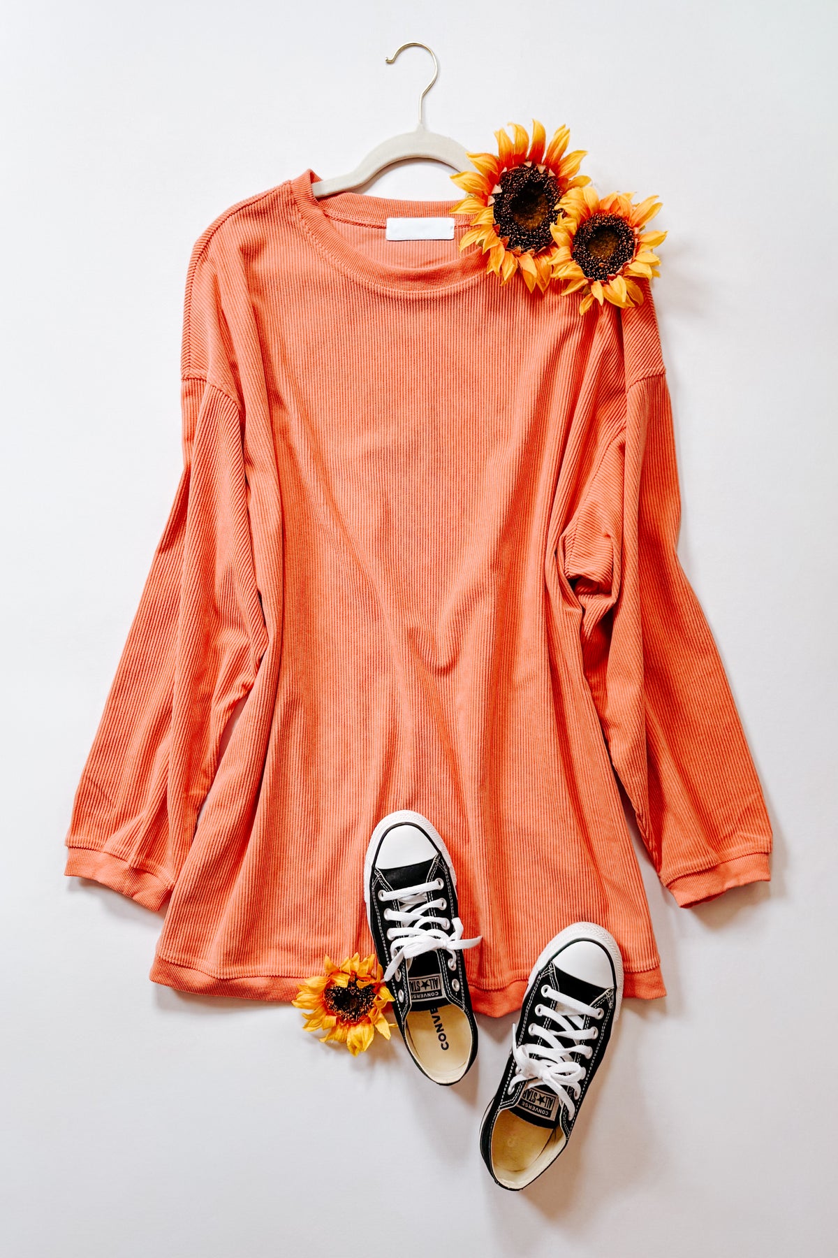 Orange corded oversized sweatshirt with ribbed trim arranged on a hanger with sunflowers and black high top sneakers for bold style